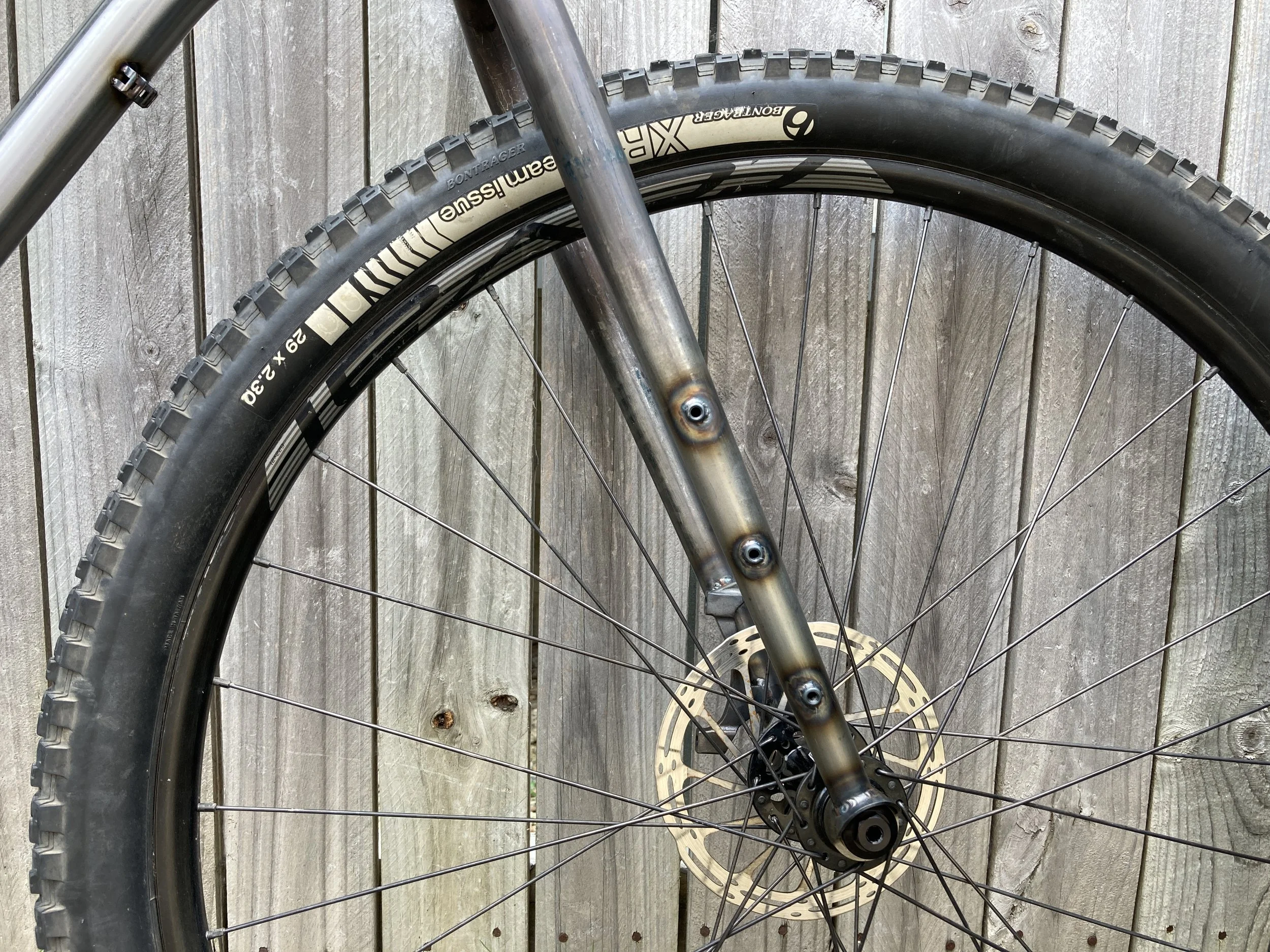 Close-up of a mountain bike front wheel with a disc brake, non-tread tire, and a part of the suspension fork, against a wooden fence background.