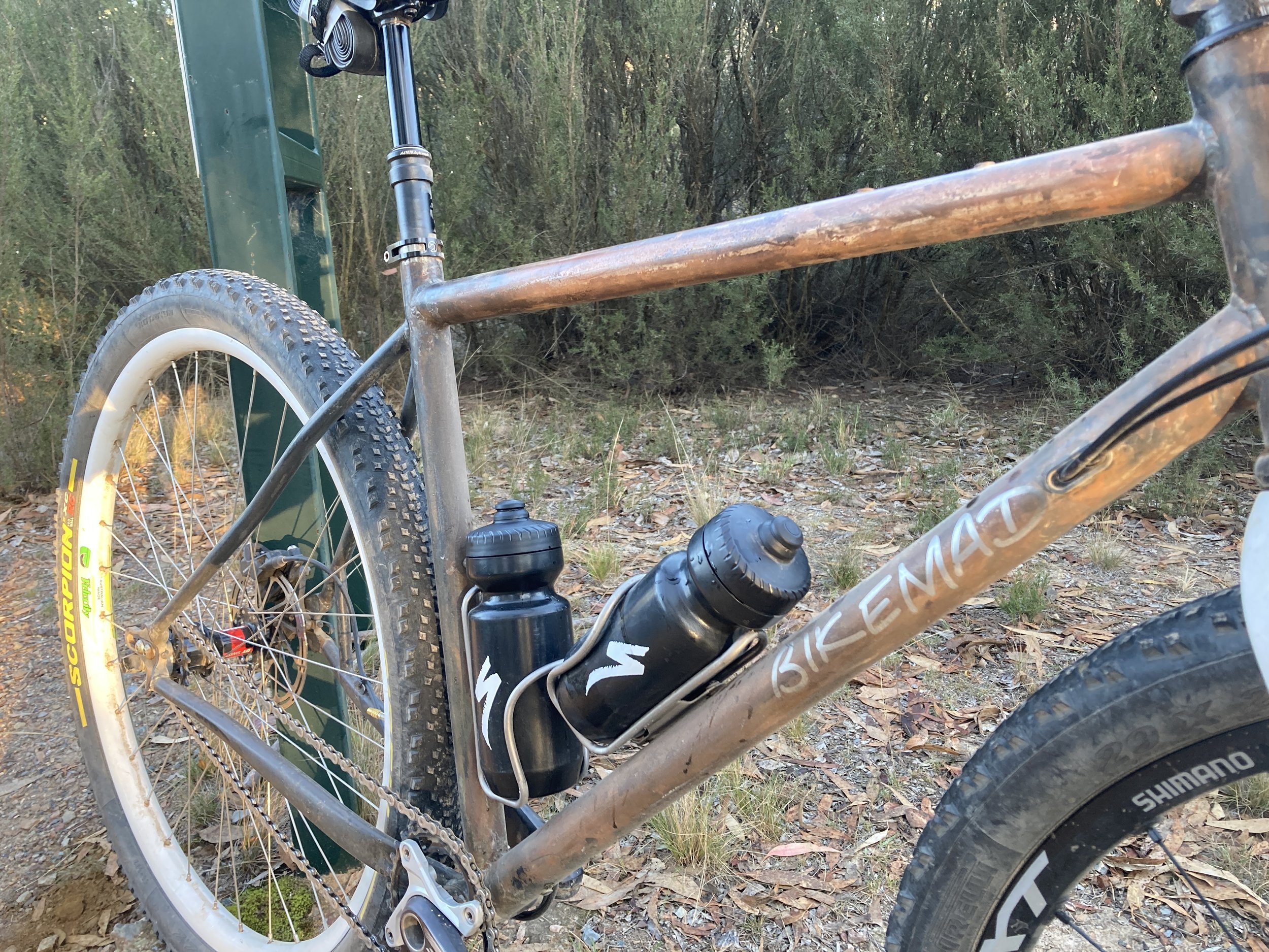 A mountain bike with a metal frame, water bottles, and wide tires, resting on a dirt trail with greenery in the background.
