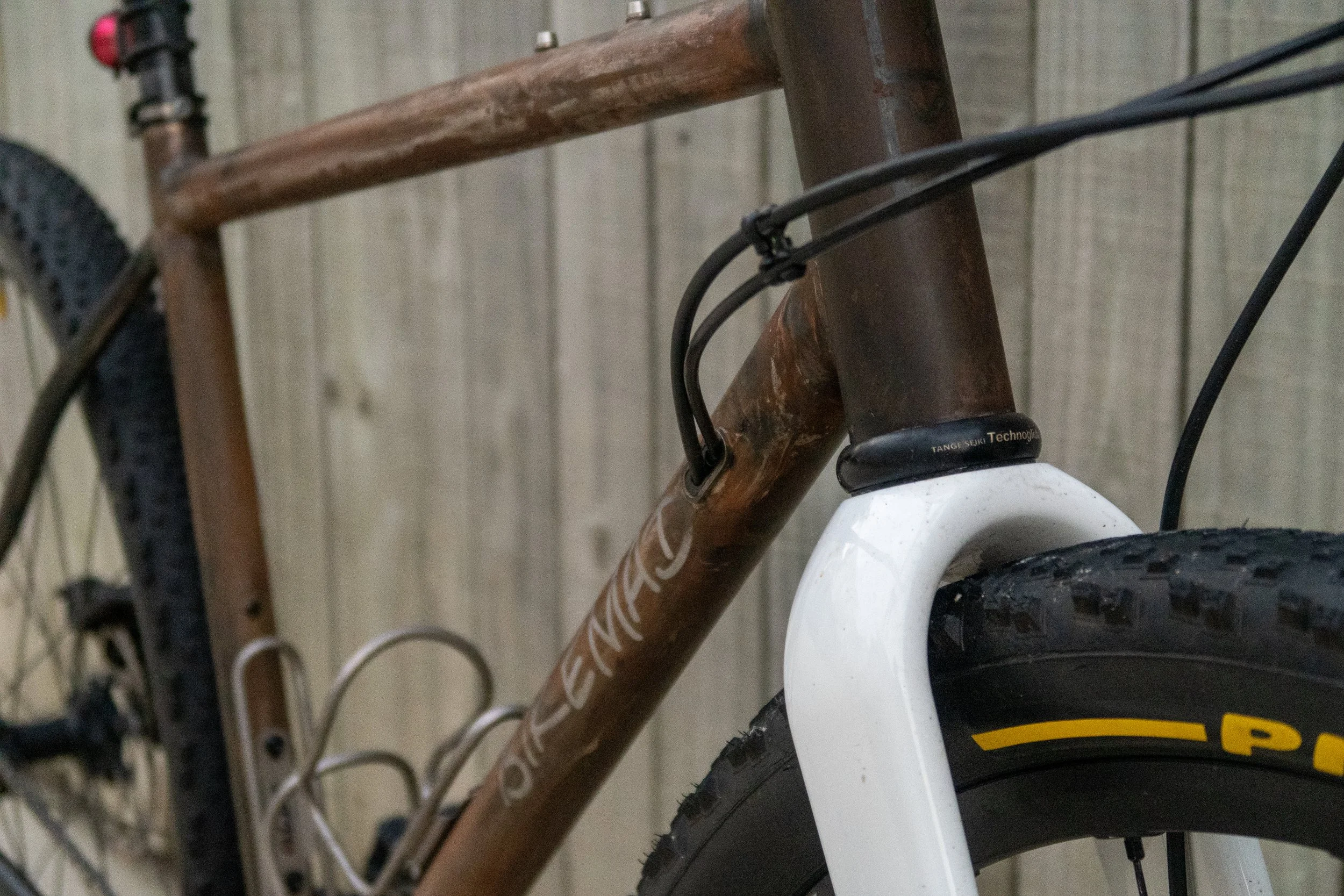 Close-up of a mountain bike's front section, showing the brown frame, black cables, white suspension fork, and front tire with a yellow logo, against a wooden background.