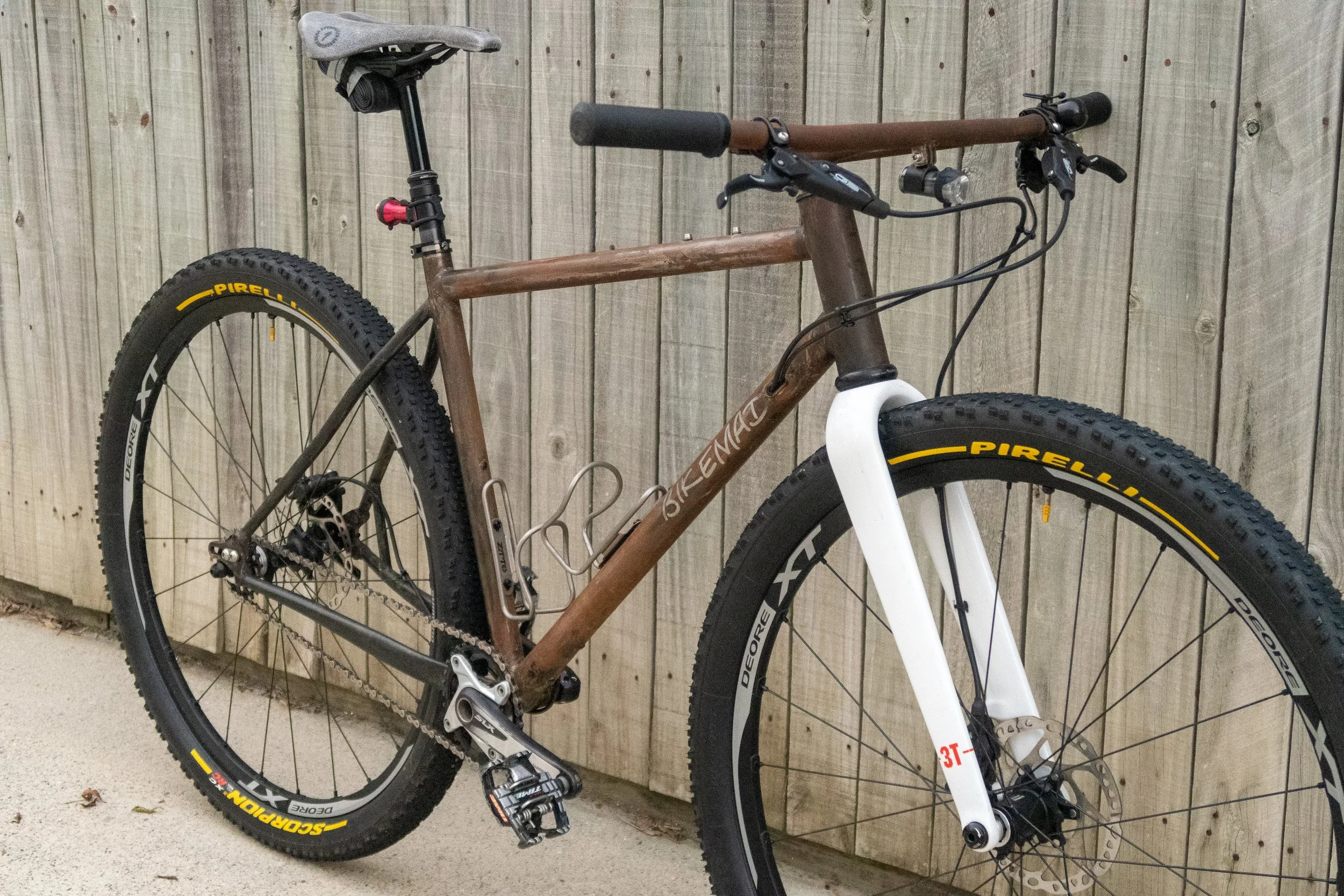 A brown Trek mountain bike leaning against a wooden fence, with black tires that have yellow Pirelli branding, a white front fork, and a black saddle.
