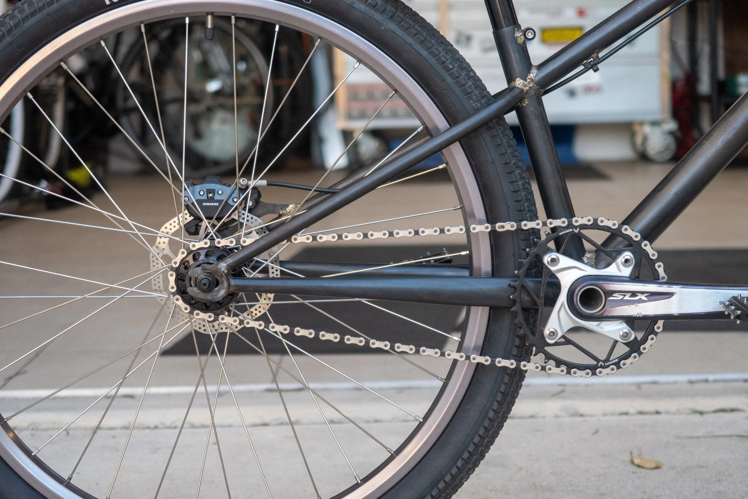 Close-up of a black bicycle's rear wheel, chain, and derailleur, with a gray rim and black tire in a garage setting.