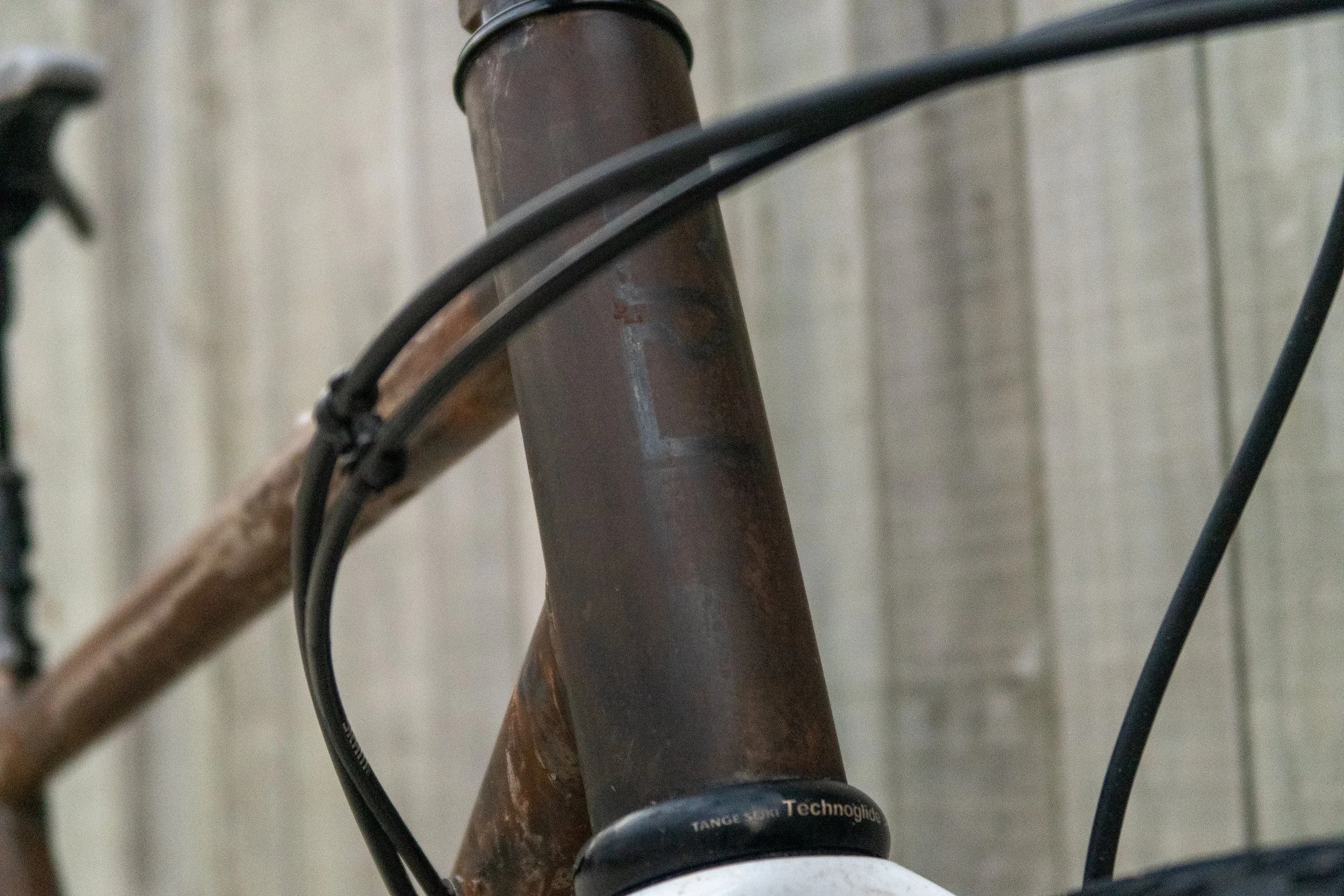 Close-up of a rusty bicycle frame with black cables attached, against a wooden wall background.