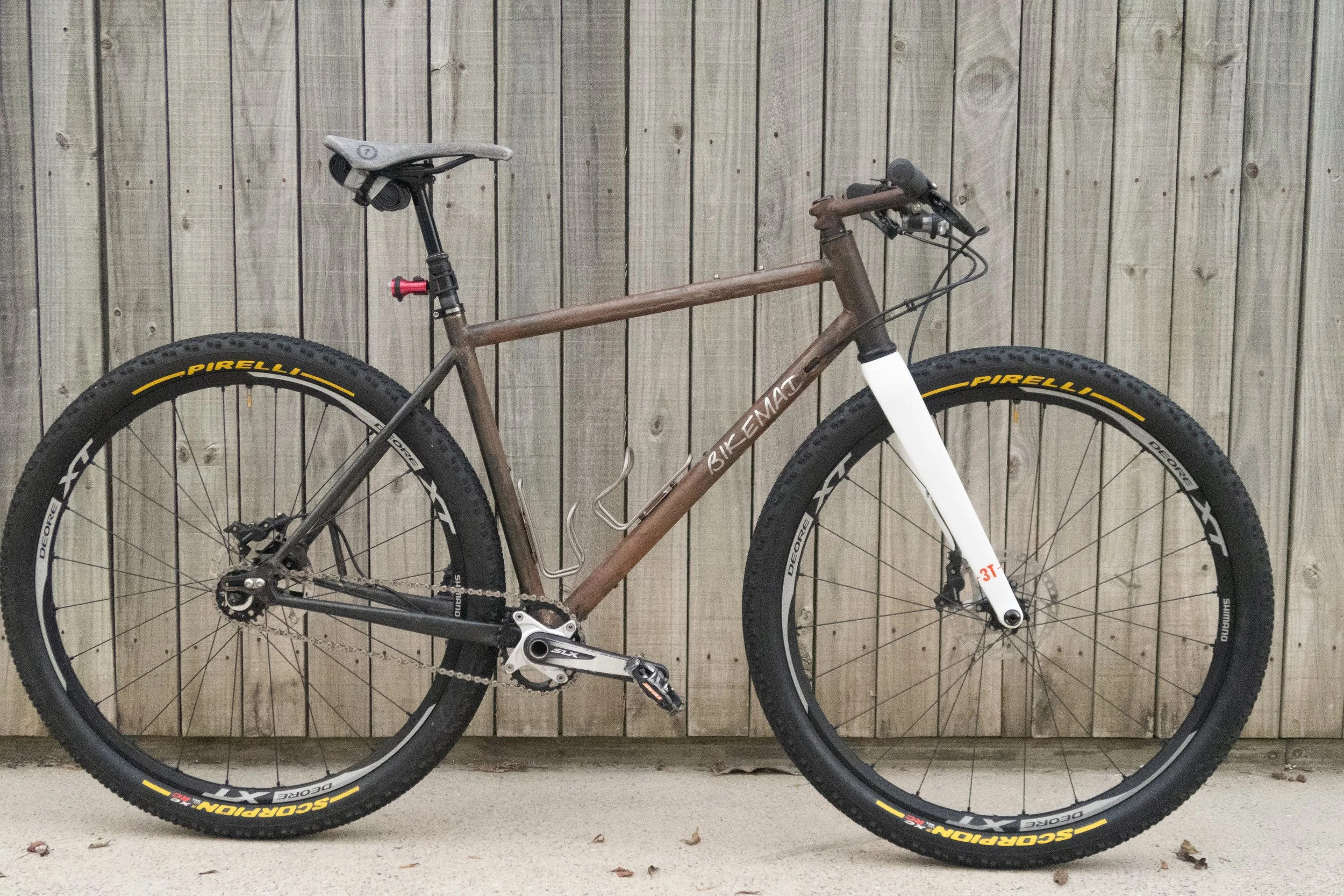 A mountain bike with a brown frame, black tires with yellow branding, and a white front fork, leaning against a wooden fence.