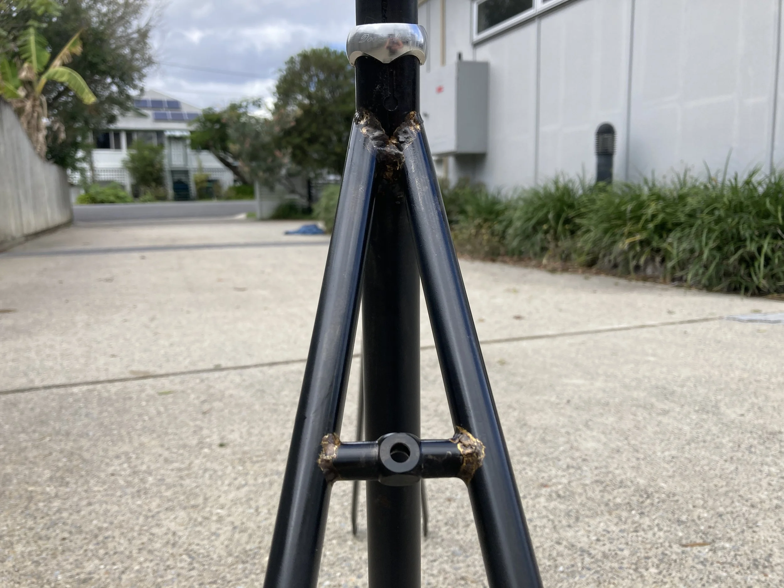 Close-up of the front part of a black bicycle fork with visible rust and damage, on a concrete driveway with houses and greenery in the background.