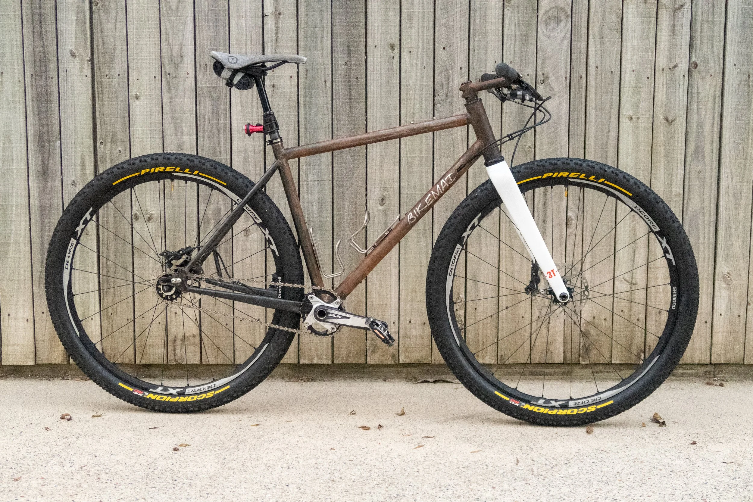 A brown mountain bike with a white front suspension fork leaning against a wooden fence.