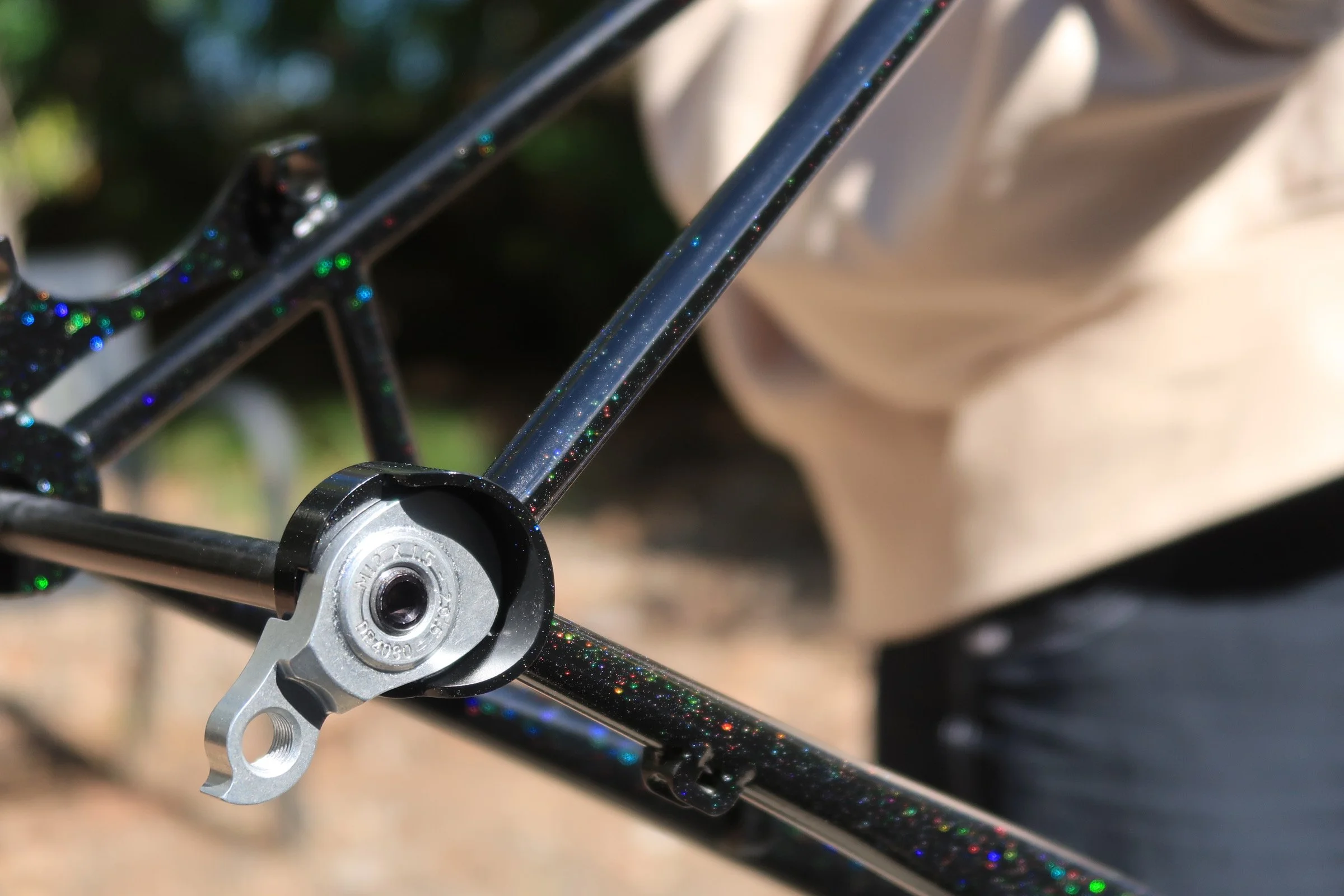 Close-up of a bicycle frame with a sparkly black finish, showing part of the rear wheel and a metal derailleur hanger, amidst a blurred background.