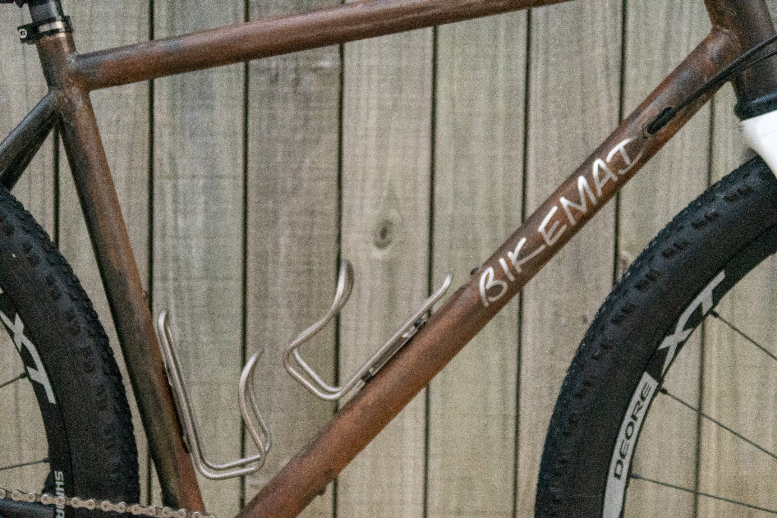 Close-up of a brown mountain bike with the words 'Bike Ematt' written on the frame, showing part of the front and rear tires and a metal water bottle holder on the frame.
