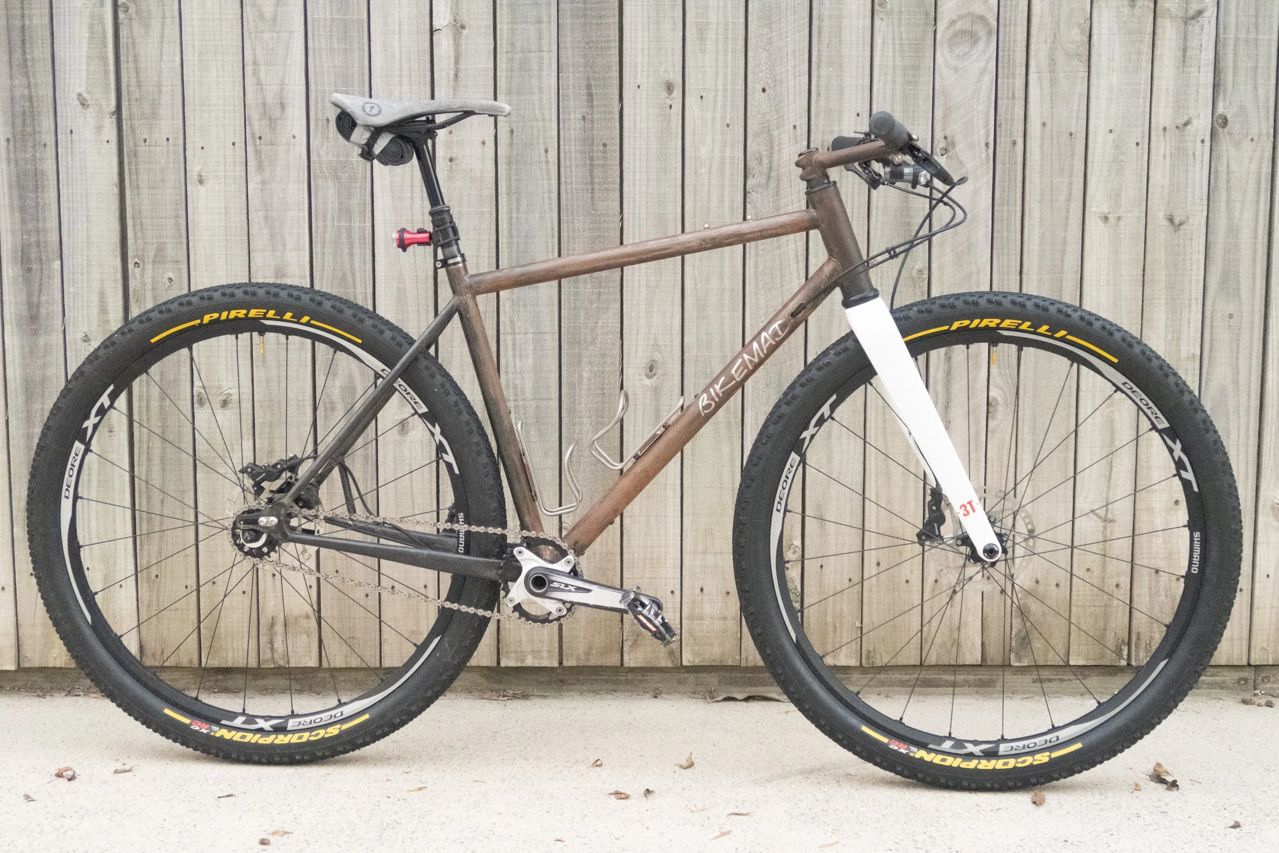 A brown and white full-suspension mountain bike with large tires, a padded seat, and a water bottle holder, leaning against a wooden fence.