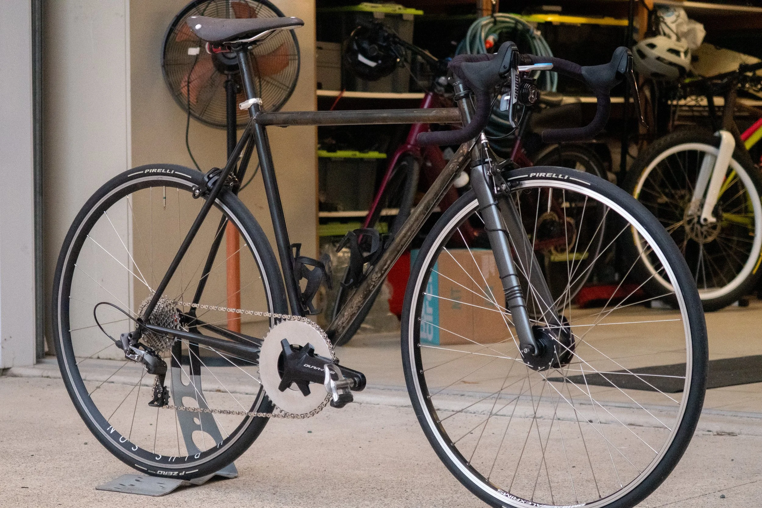 A black road bicycle with drop handlebars, a gear system, and Pirelli tires, leaning on a kickstand in a garage.
