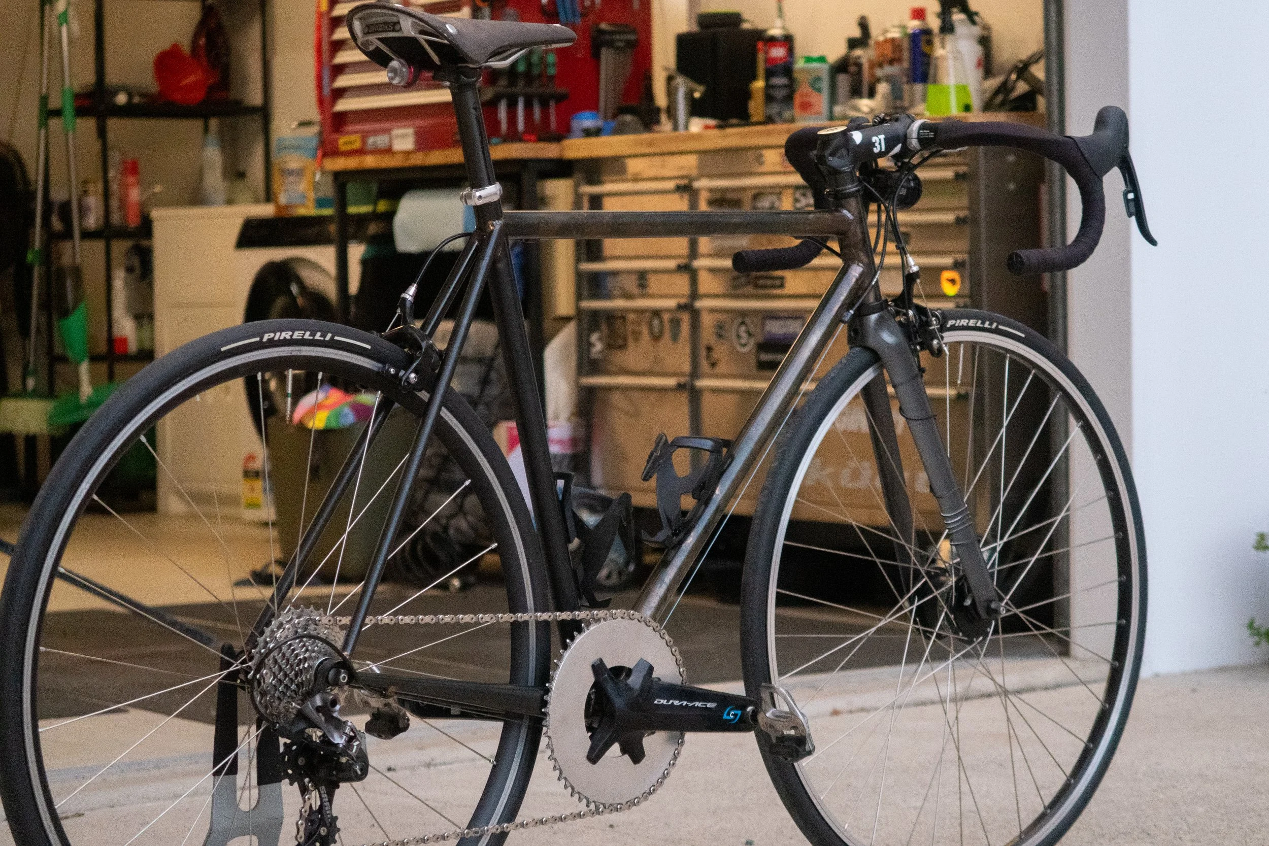 A black and silver road bicycle with Pirelli tires, parked in a garage with various tools and storage shelves in the background.