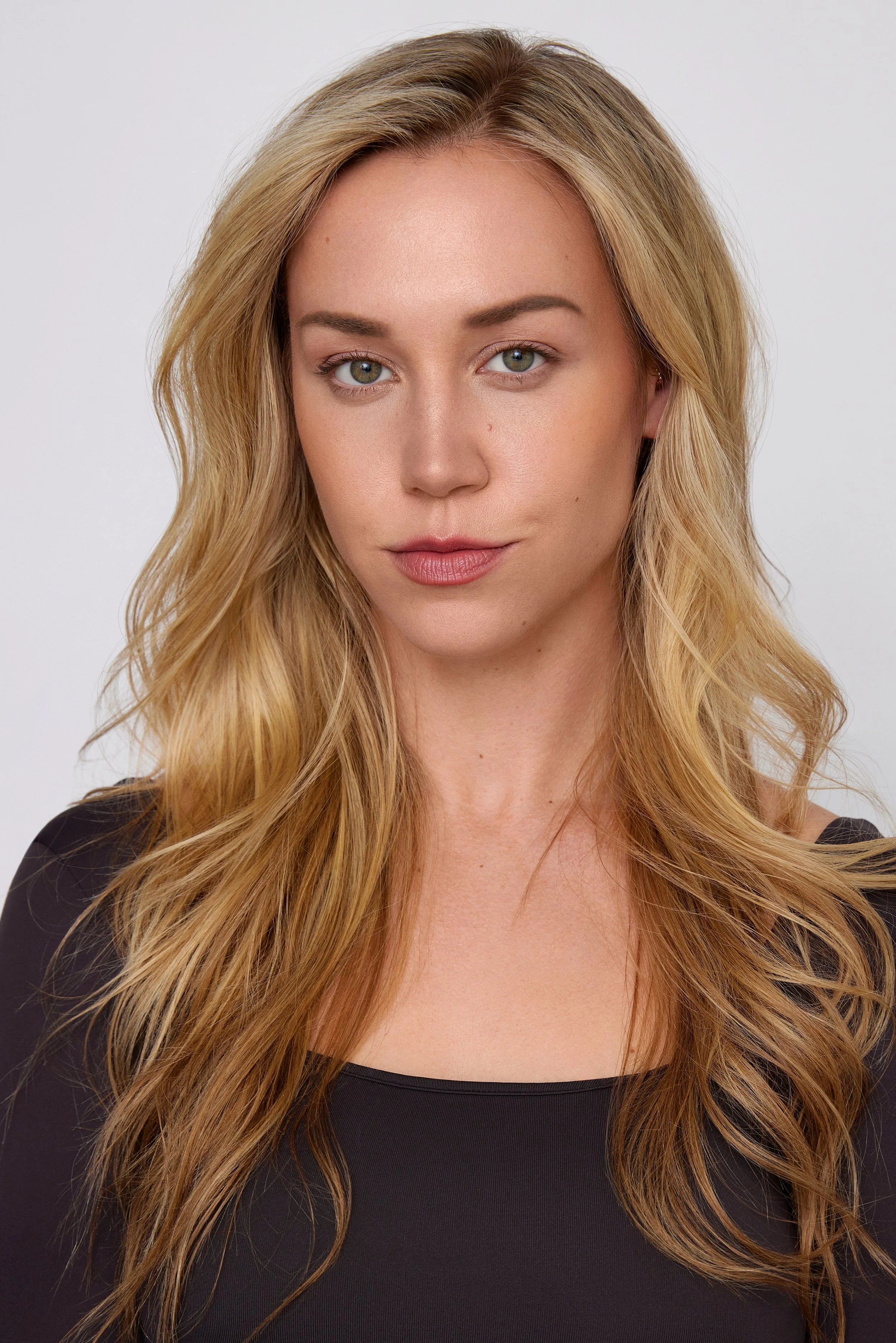 Close-up portrait of a young woman with long, wavy blonde hair, blue eyes, wearing a black top, against a plain white background.