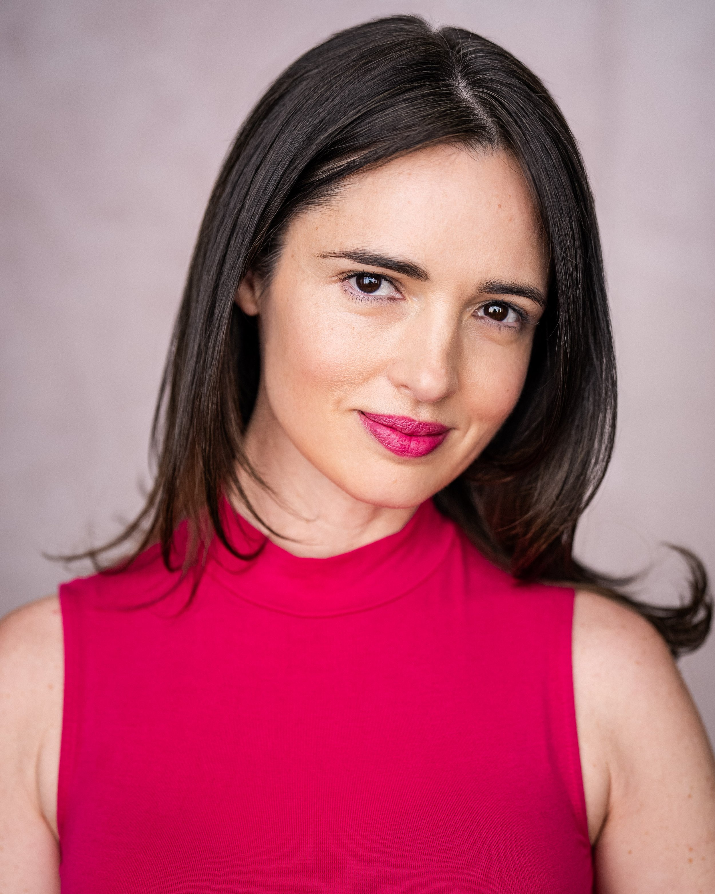 Portrait of a woman with dark hair wearing a pink sleeveless top, smiling softly, with a neutral gray background.