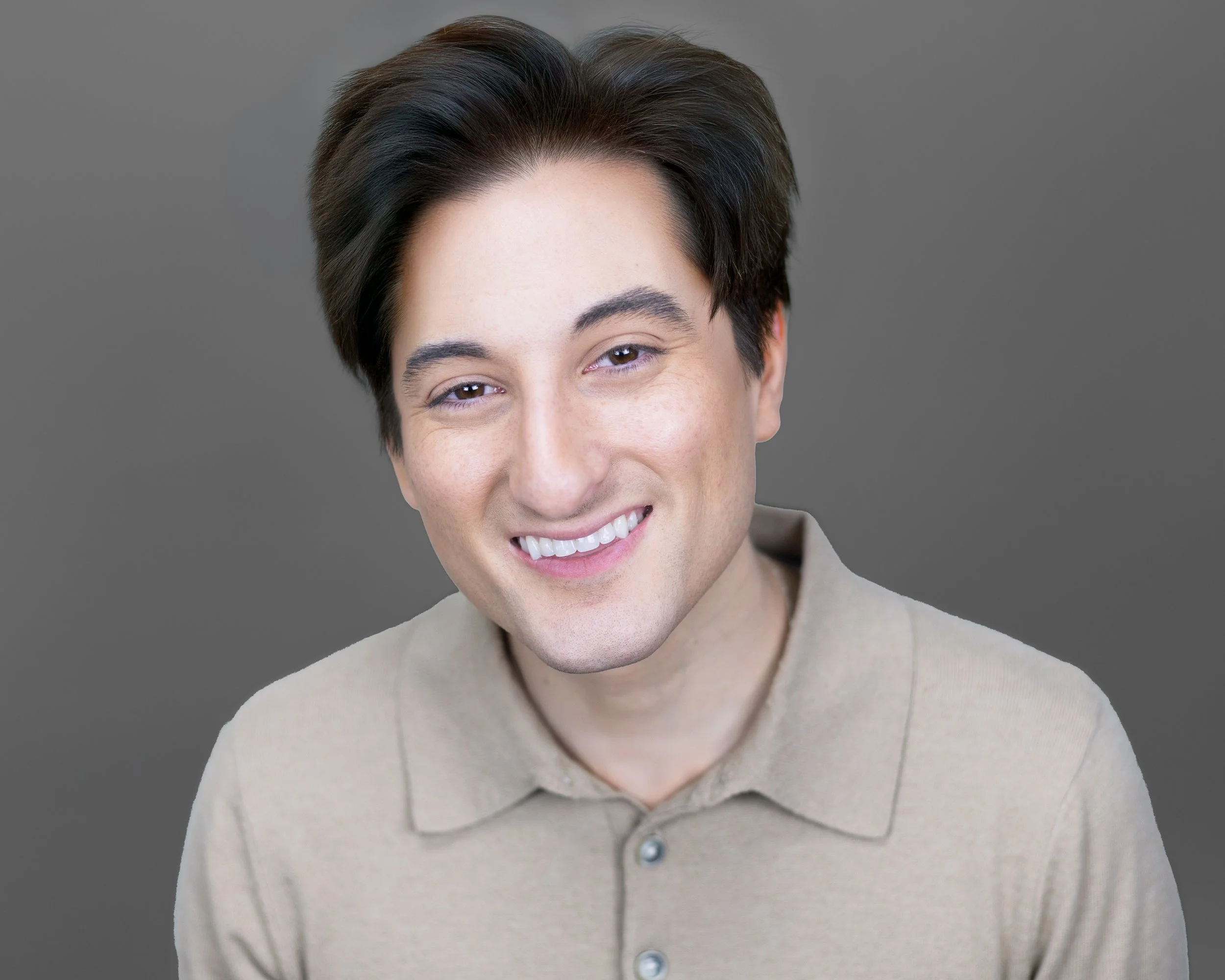 Portrait of a young man with dark brown hair, smiling, wearing a beige collared shirt, against a gray background.