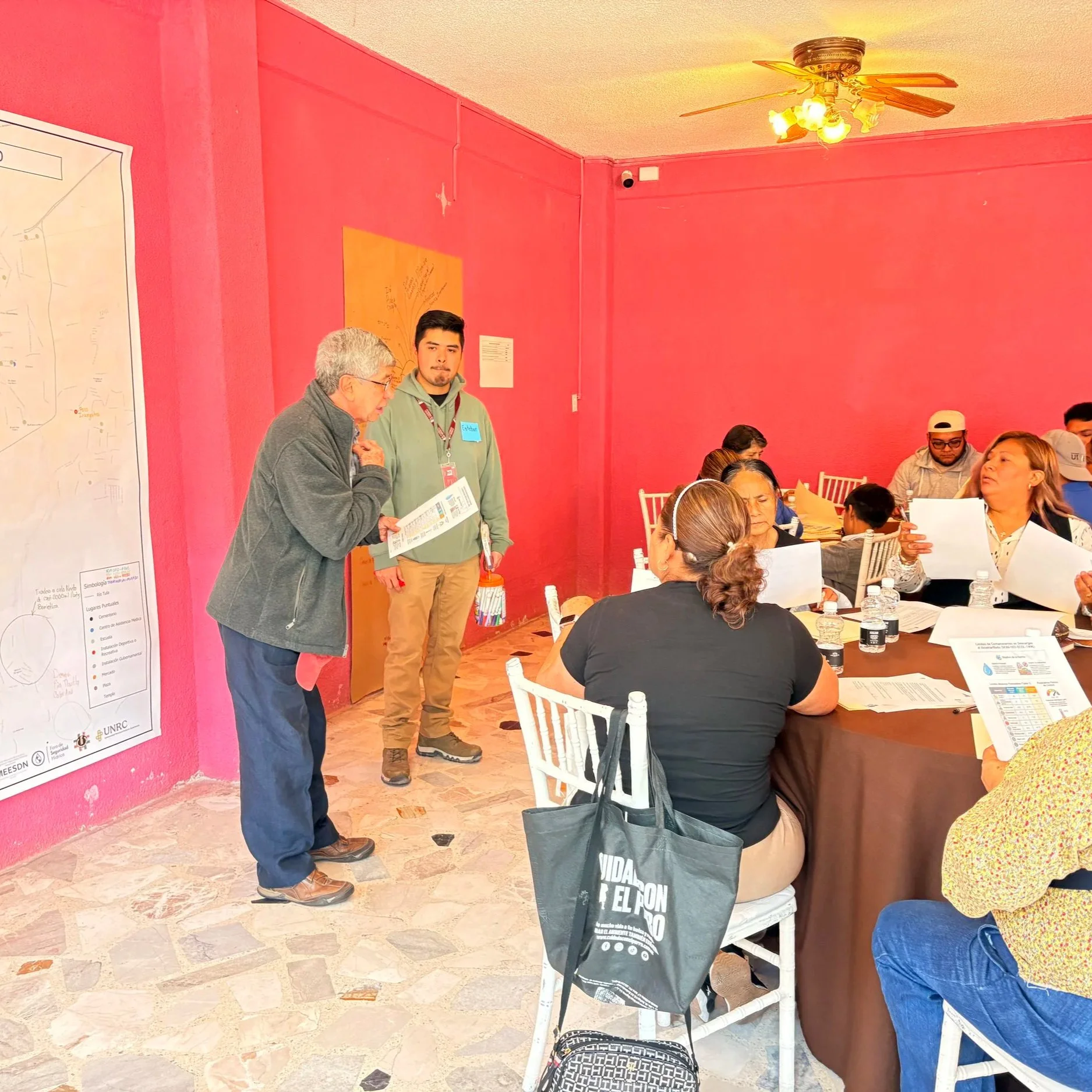 Reunión de varias personas en una mesa, mientras dos hombres en el frente discuten y revisan papeles, en una habitación con paredes rosadas y piso de piedra, algunos usan gafas y llevan documentos en mano.