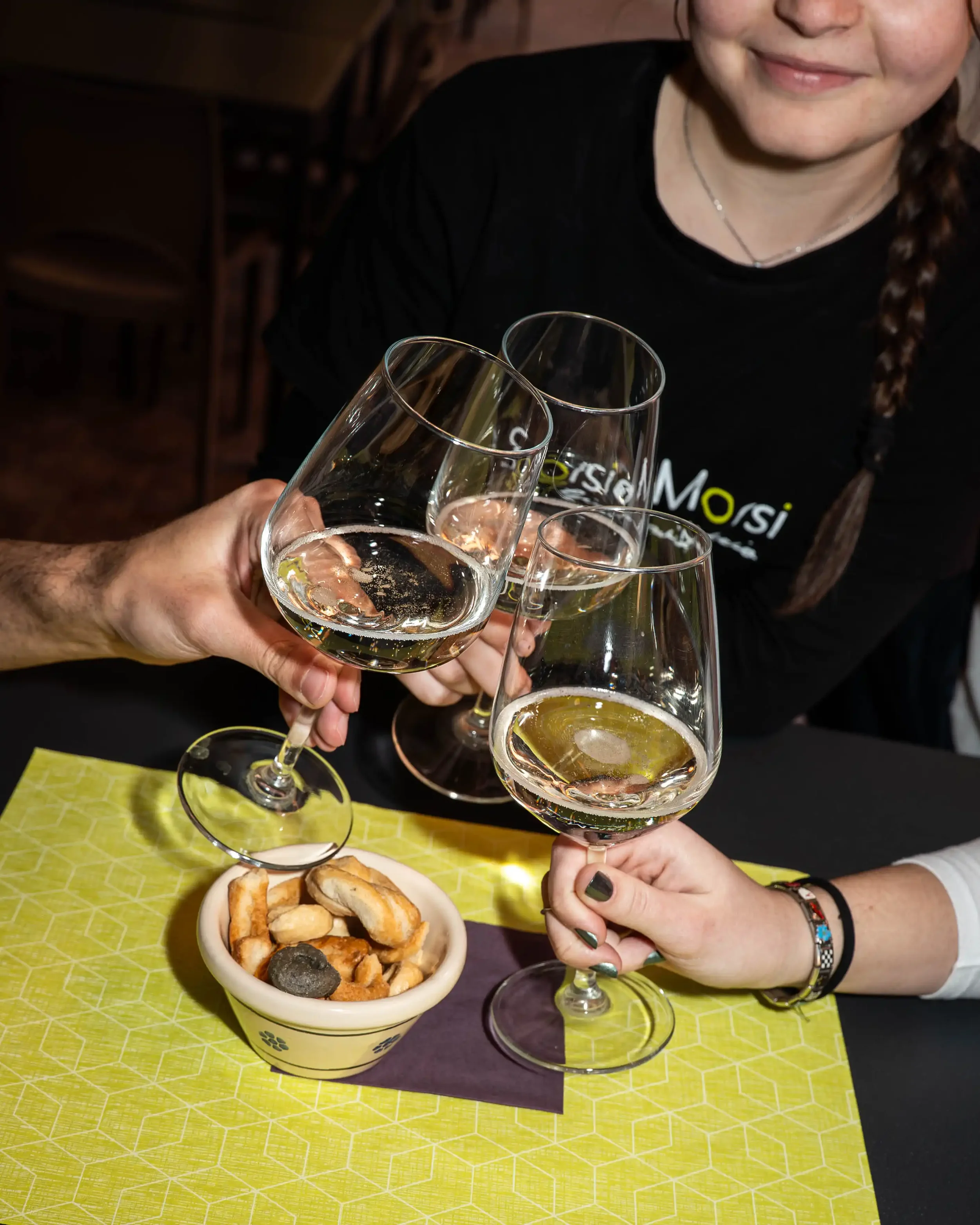 Brindisi di bicchieri di vino bianco trasparente durante un brindisi tra persone, con un piatto di biscotti sul tavolo. Una donna sorridente indossa una maglietta nera e bracciali, e tiene in mano un bicchiere di vino. Sul tavolo una tovaglia gialla con motivo geometrico.