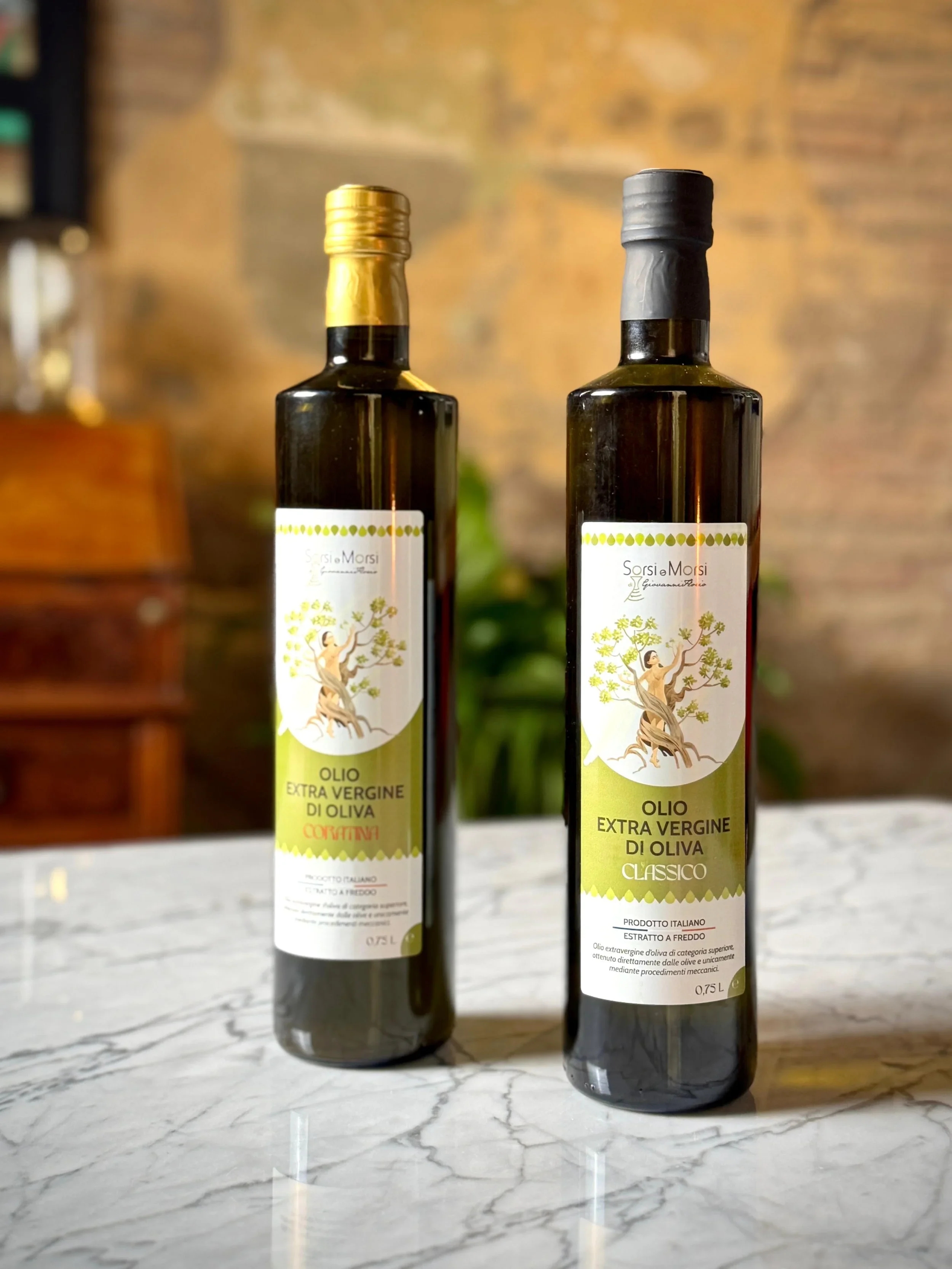 Bottiglie di olio extravergine di oliva con etichette decorative, posizionate su un tavolo di marmo con sfondo di mattoni e piante verde in cucina.