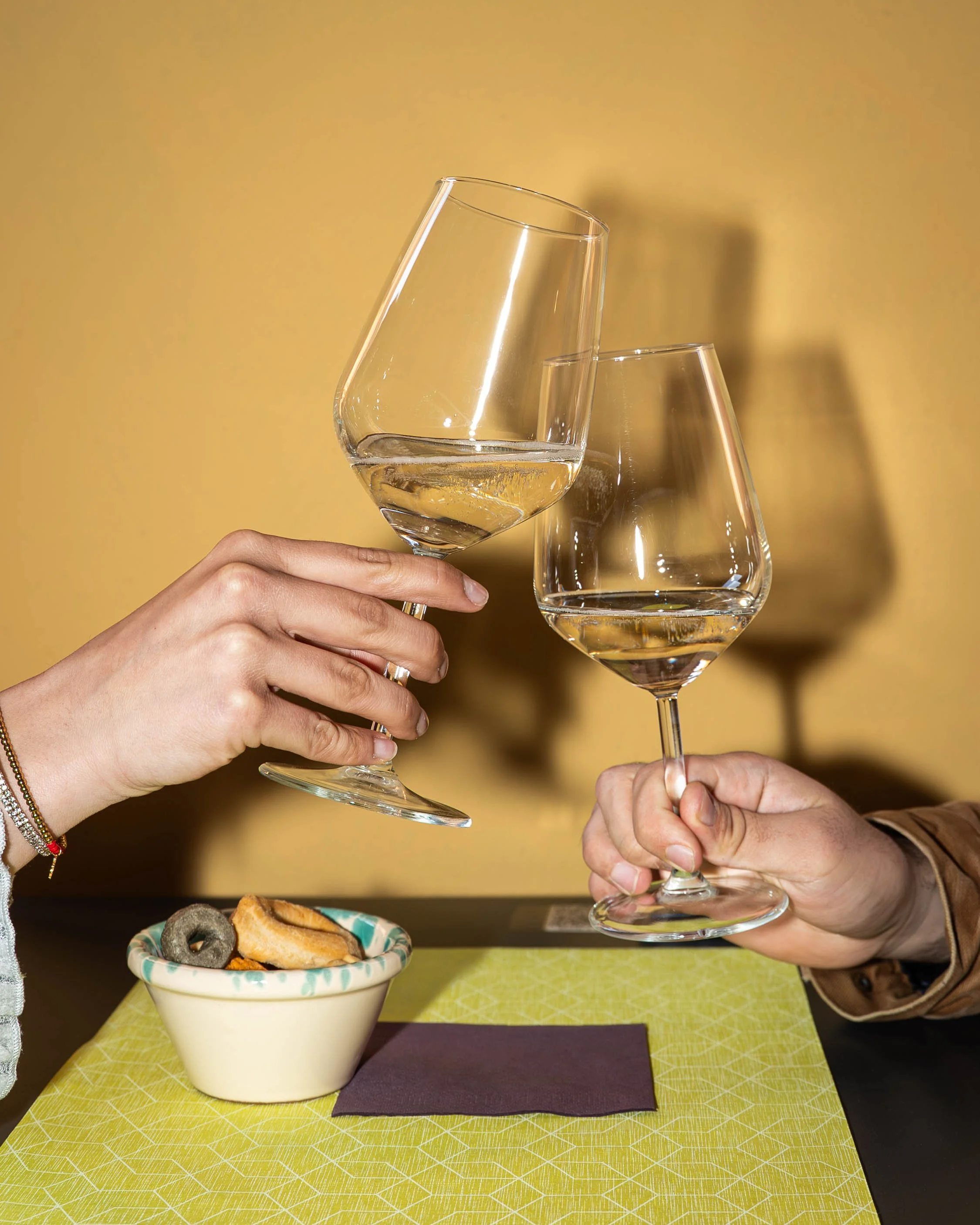 Due persone che brindano con calici di vino bianco, con un piatto di biscotti e snack sul tavolo.