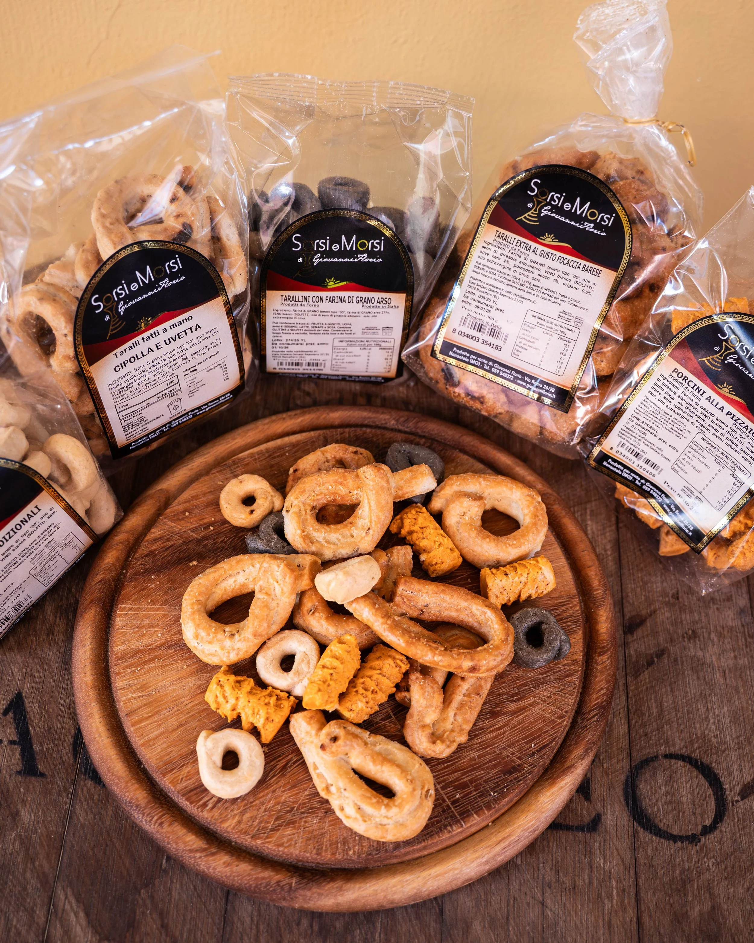 Varie tipi di taralli artigianali confezionati in sacchetti di plastica con etichette, disposti su un piatto di legno, con alcuni taralli aperti e altri chiusi, sul tavolo in legno.