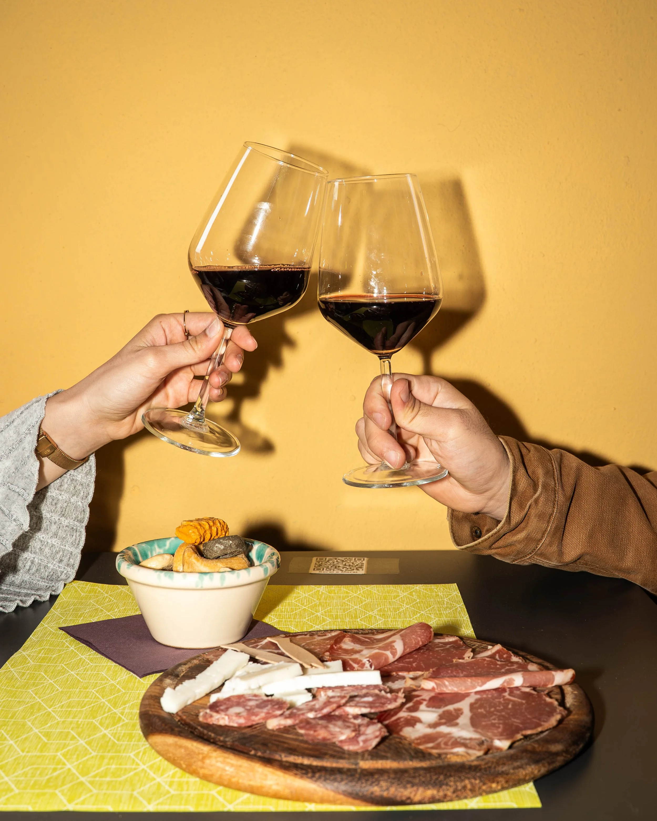 Bicchiere di vino rosso, concentrato, tenuto da una mano, in una parete color giallo. Sulla tavola ci sono formaggi e affettati, presso Sorsi e Morsi a Nonantola.