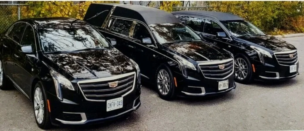 Three black Cadillac vehicles parked side by side on a street with autumn foliage in the background.
