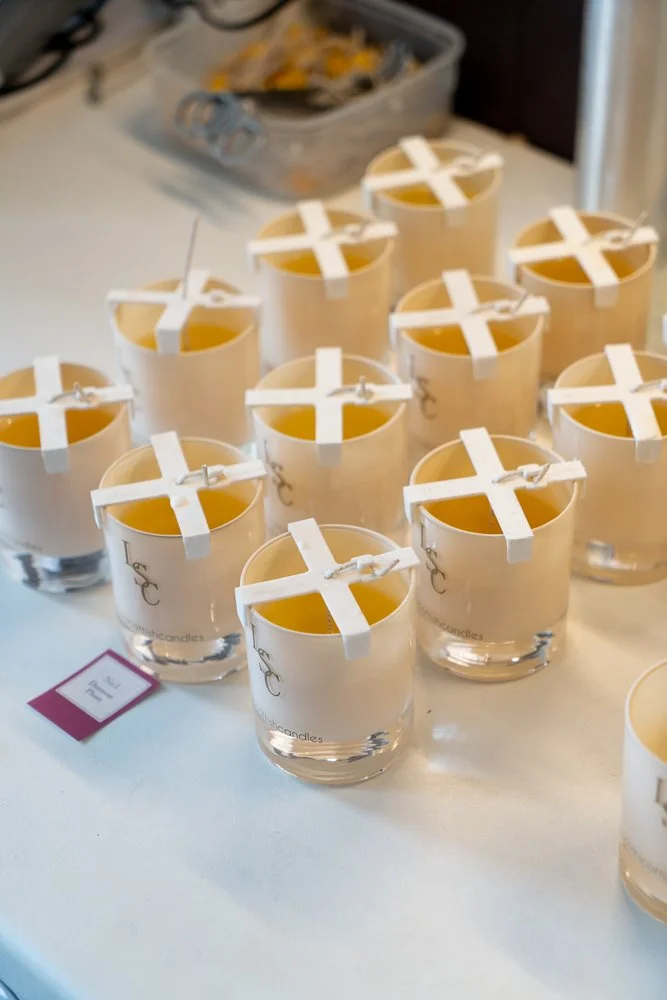 Multiple unlit candles in glass holders with wicks supported by plastic clips on a table.