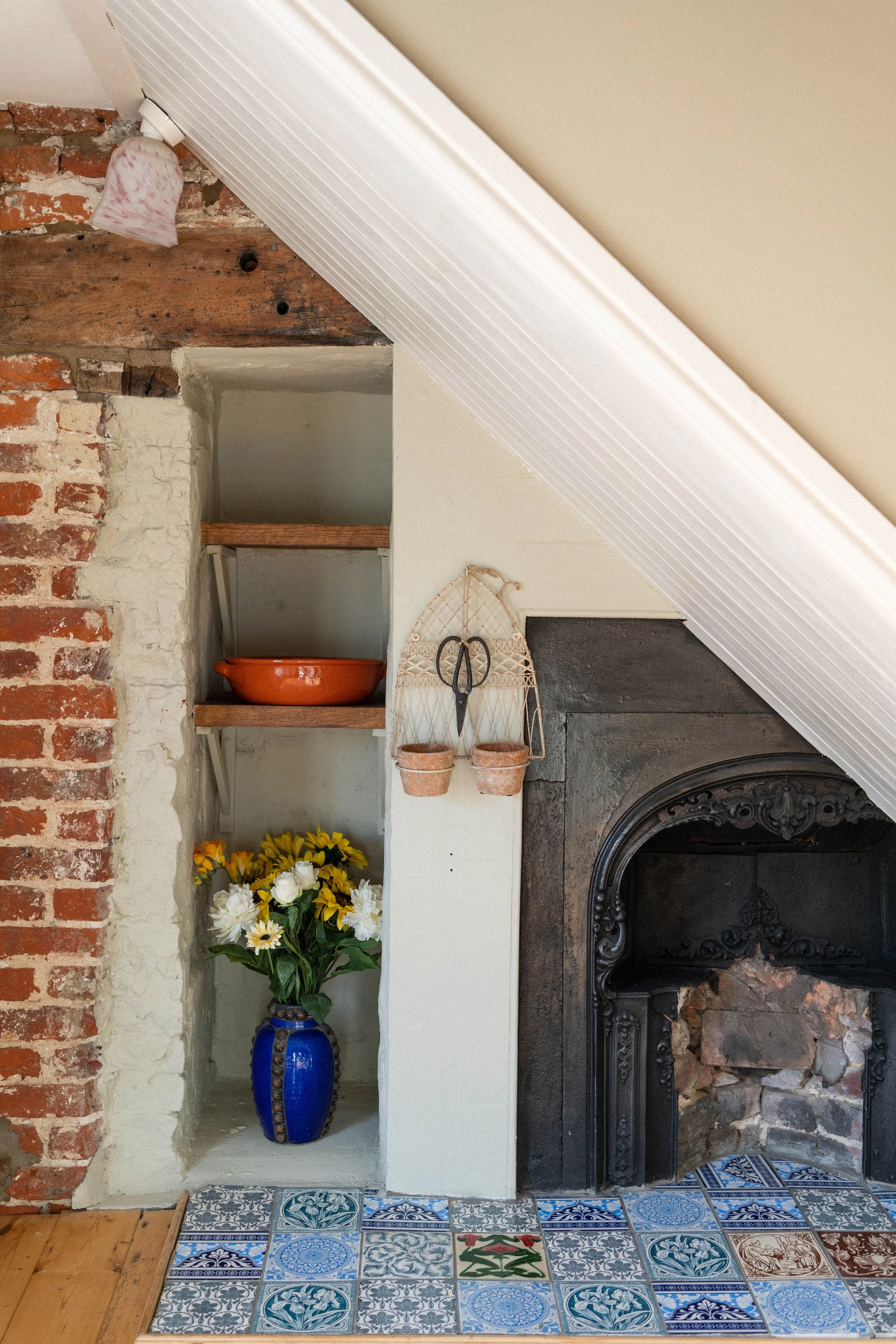 Cozy room corner with exposed brick wall, shelf holding a clay pot, vase with flowers, ornate fireplace, and decorative tiles.