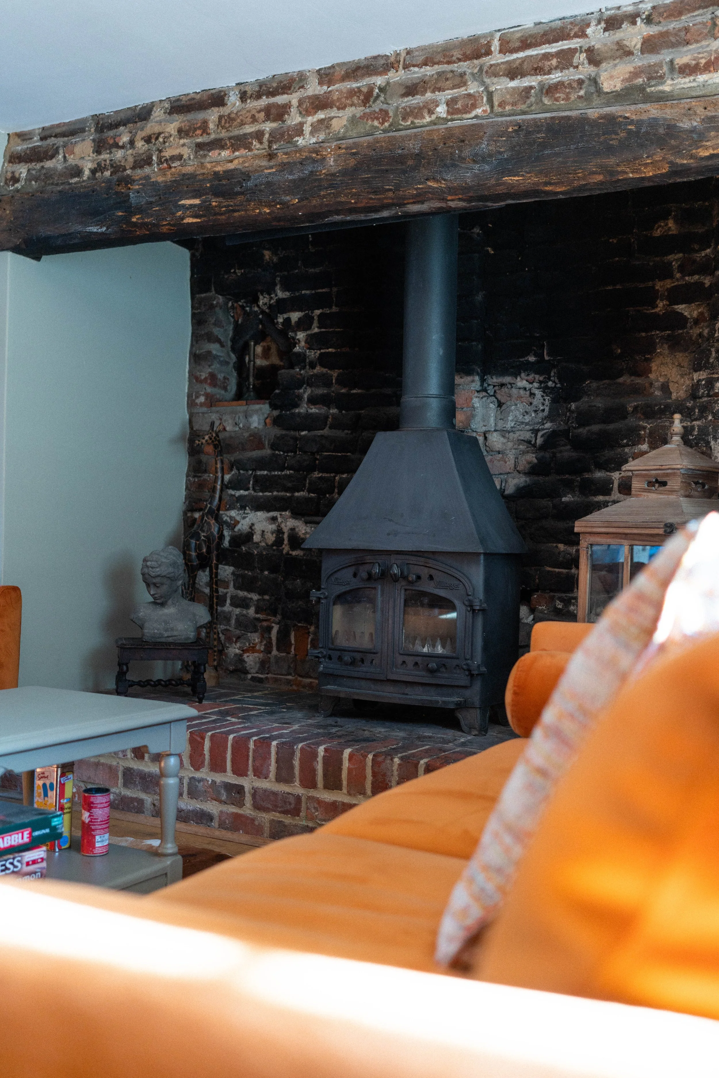 Cozy room with a brick and stone fireplace, featuring a black wood burning stove. There's a wooden beam above the fireplace, a bust sculpture on a small table, a large lantern, and a glimpse of an orange sofa and a table with board games and canned f