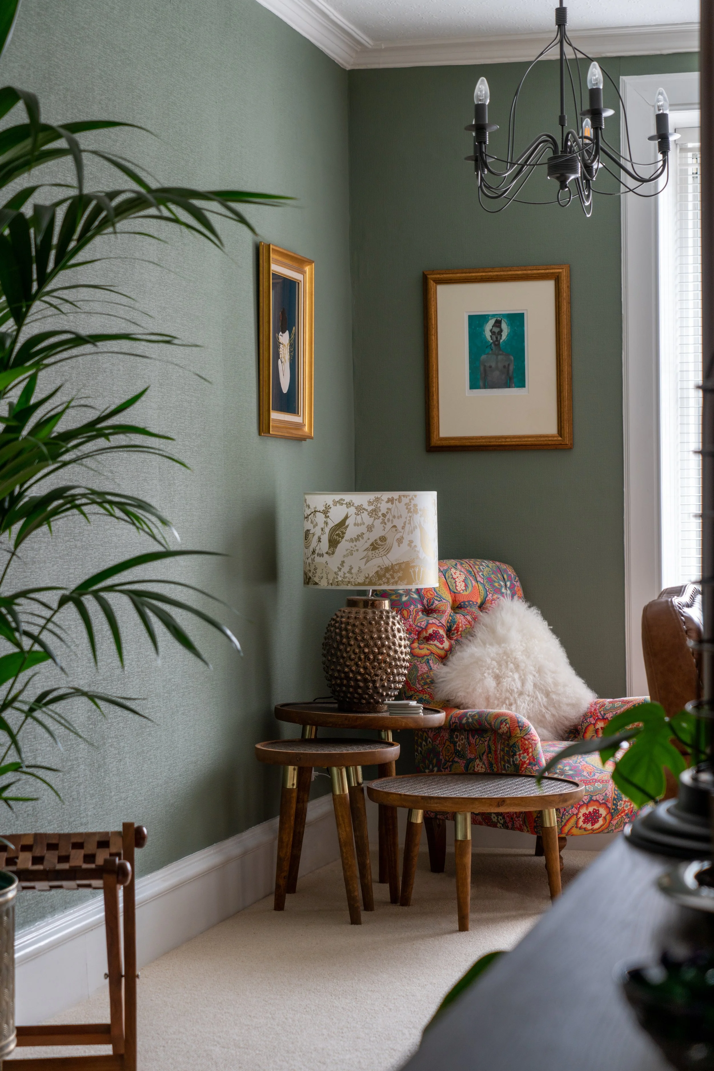 Cozy living room corner with green walls, a colorful upholstered armchair, wooden side tables, potted plants, and framed artwork. A decorative lamp sits on the table beside the chair, under a modern chandelier.