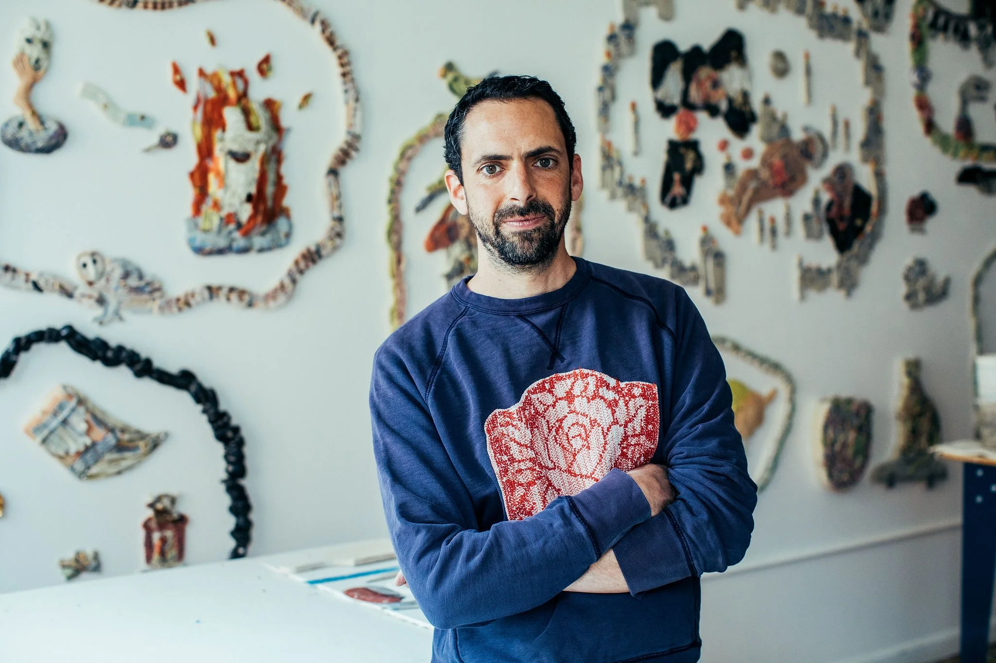 Man with dark hair, beard, and blue eyes wearing a navy sweatshirt is standing in front of an art installation made of various textured materials on a white wall. Features mounted ceramic artwork resembling animals and abstract shapes. Judd Schiffman