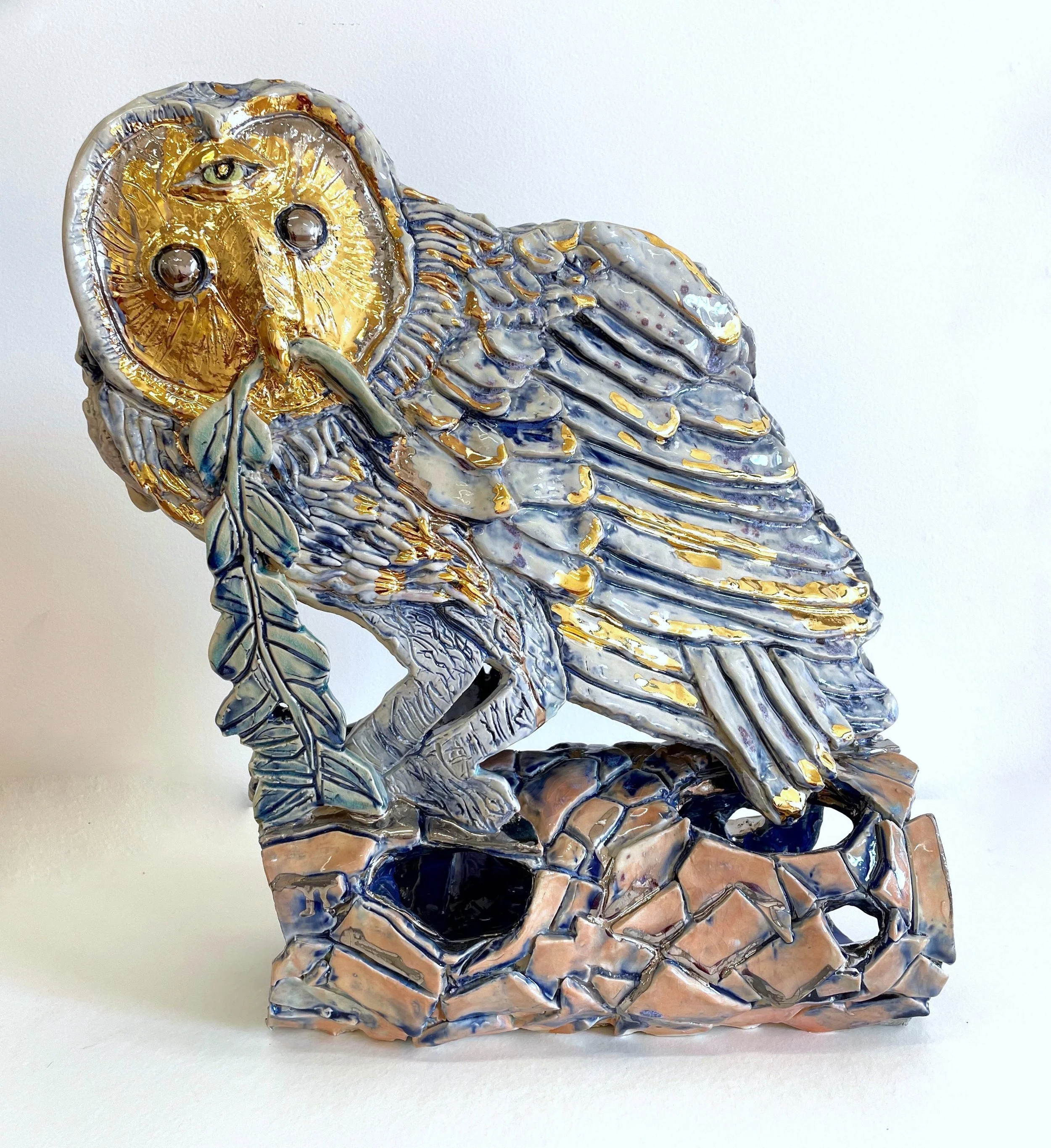 A decorative ceramic sculpture of an owl perched on a branch with a feather in its beak, featuring gold and blue accents.