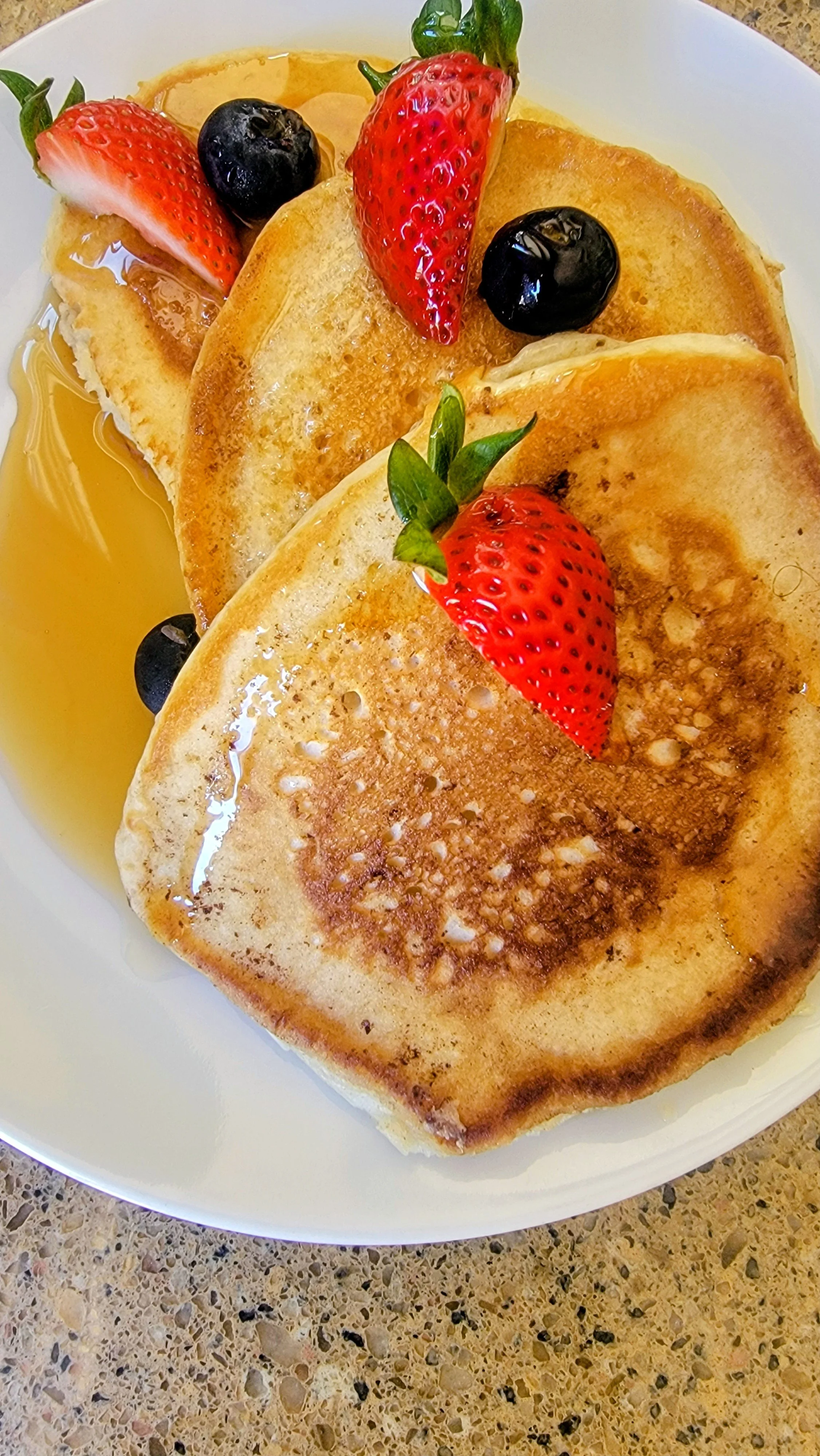 A plate of two golden-brown pancakes topped with fresh strawberries and blueberries, with syrup drizzled on the side.