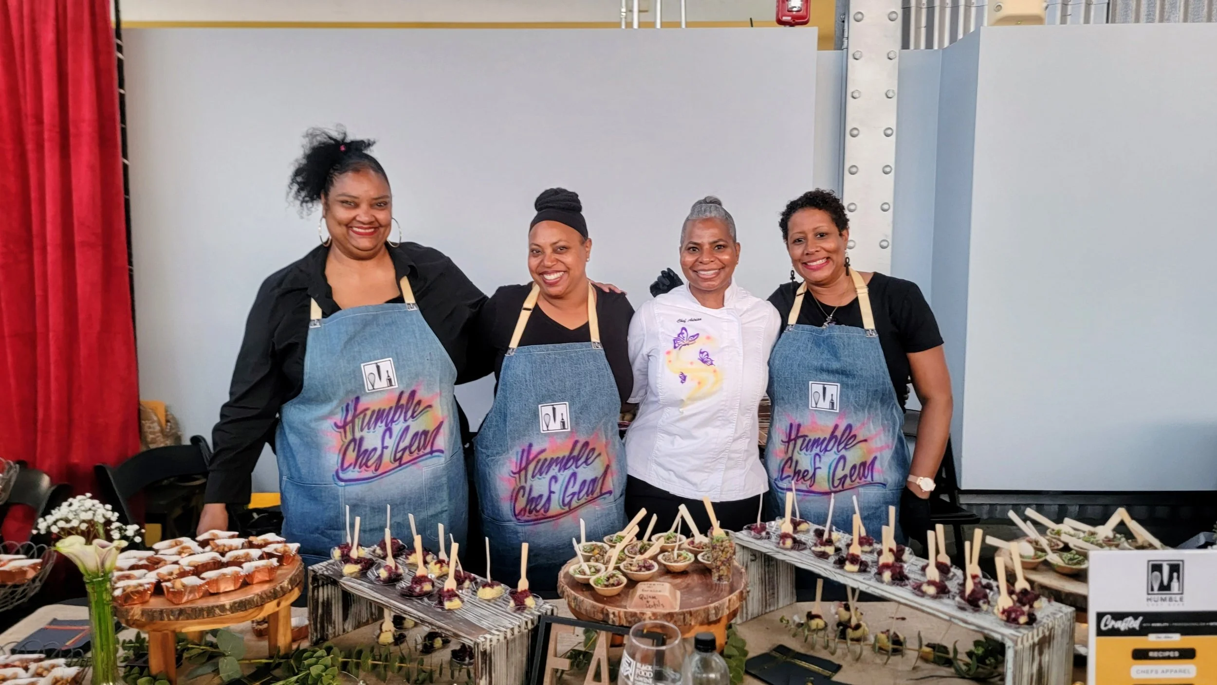 Four women standing behind a table filled with desserts, wearing aprons that say 'Humble Chef Gear,' at a culinary event.