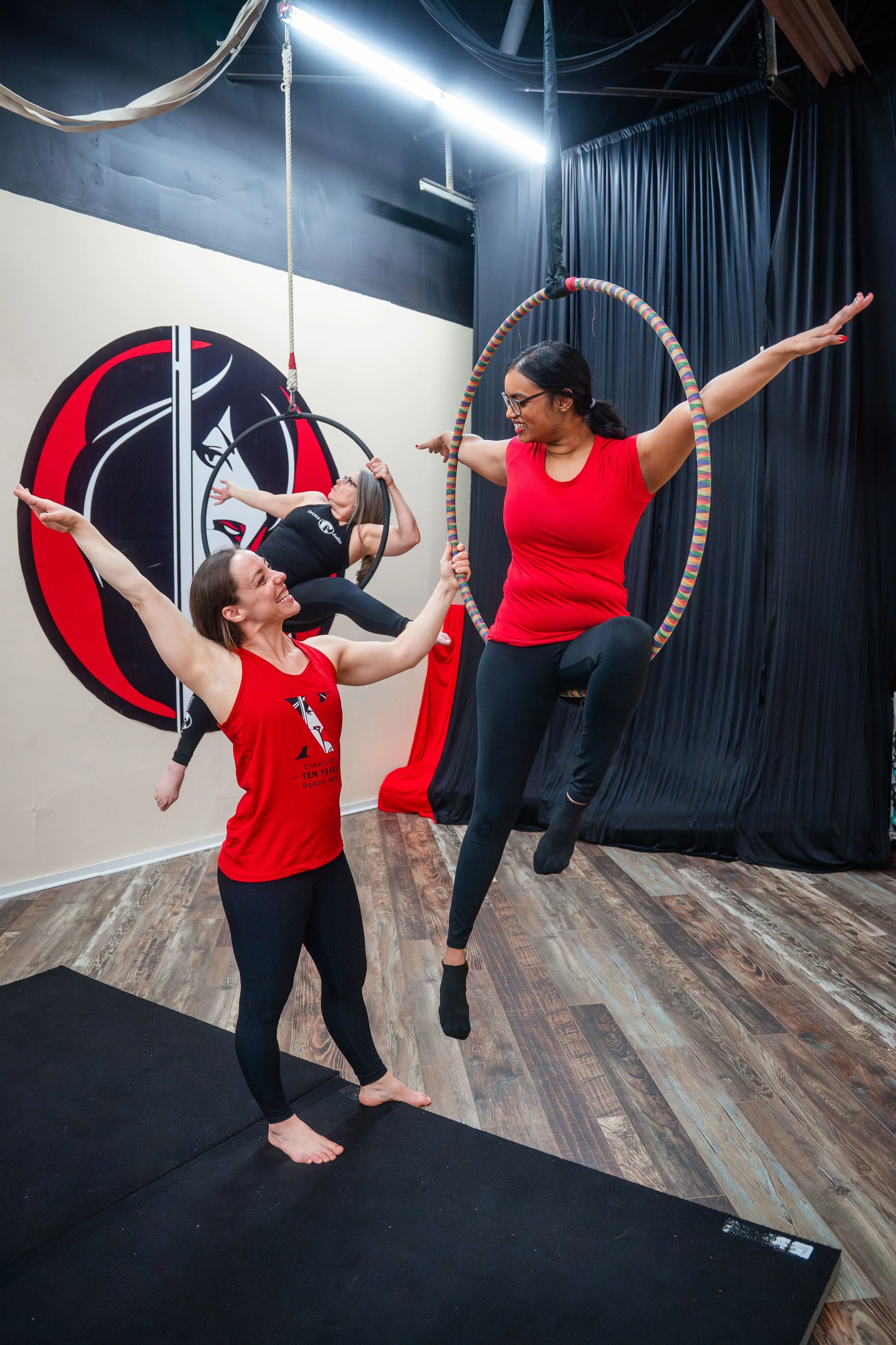 Three women are practicing aerial hoop and silks in a gym with a large logo of a clown face on the wall. One woman is standing on a mat, smiling, holding an aerial hoop, while another is sitting in a hoop hanging from the ceiling, and the third is hanging from silks in the background.