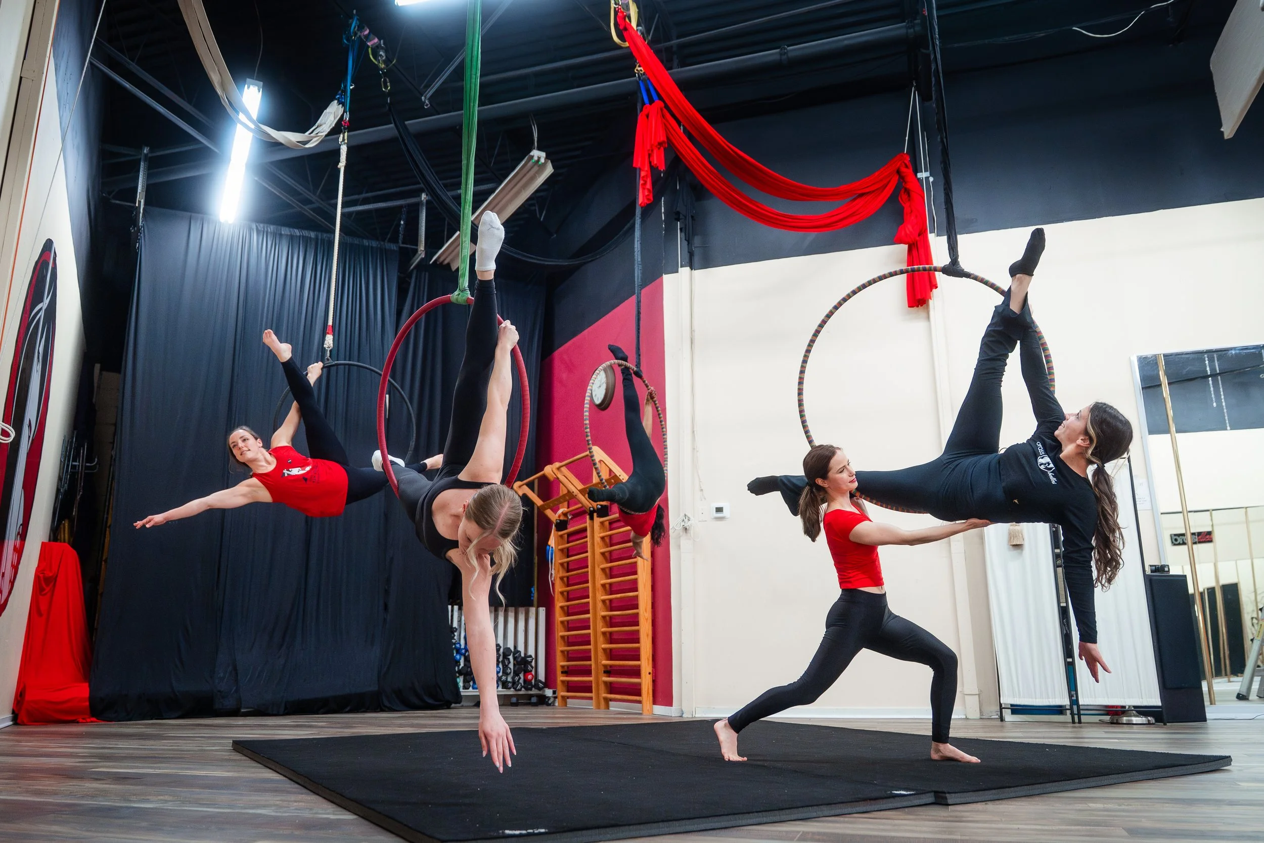Four women practicing aerial silk and acrobatics in a gym, with two hanging from red hoops and one on the ground assisting, in a fitness studio with black, red, and cream walls.