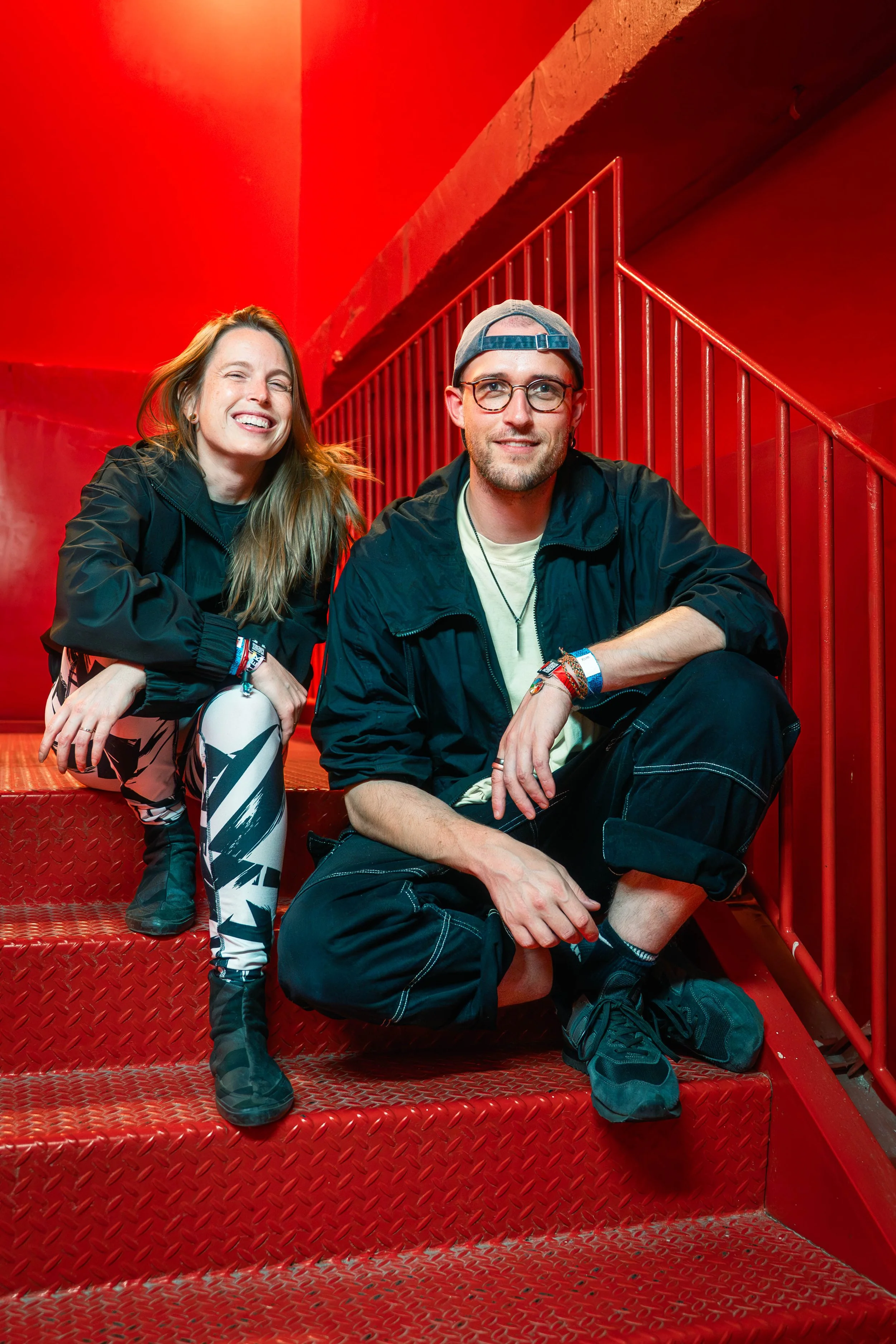 A young woman and a young man sitting on red metal stairs, smiling, with a red background and metal railing, wearing casual clothes and accessories.