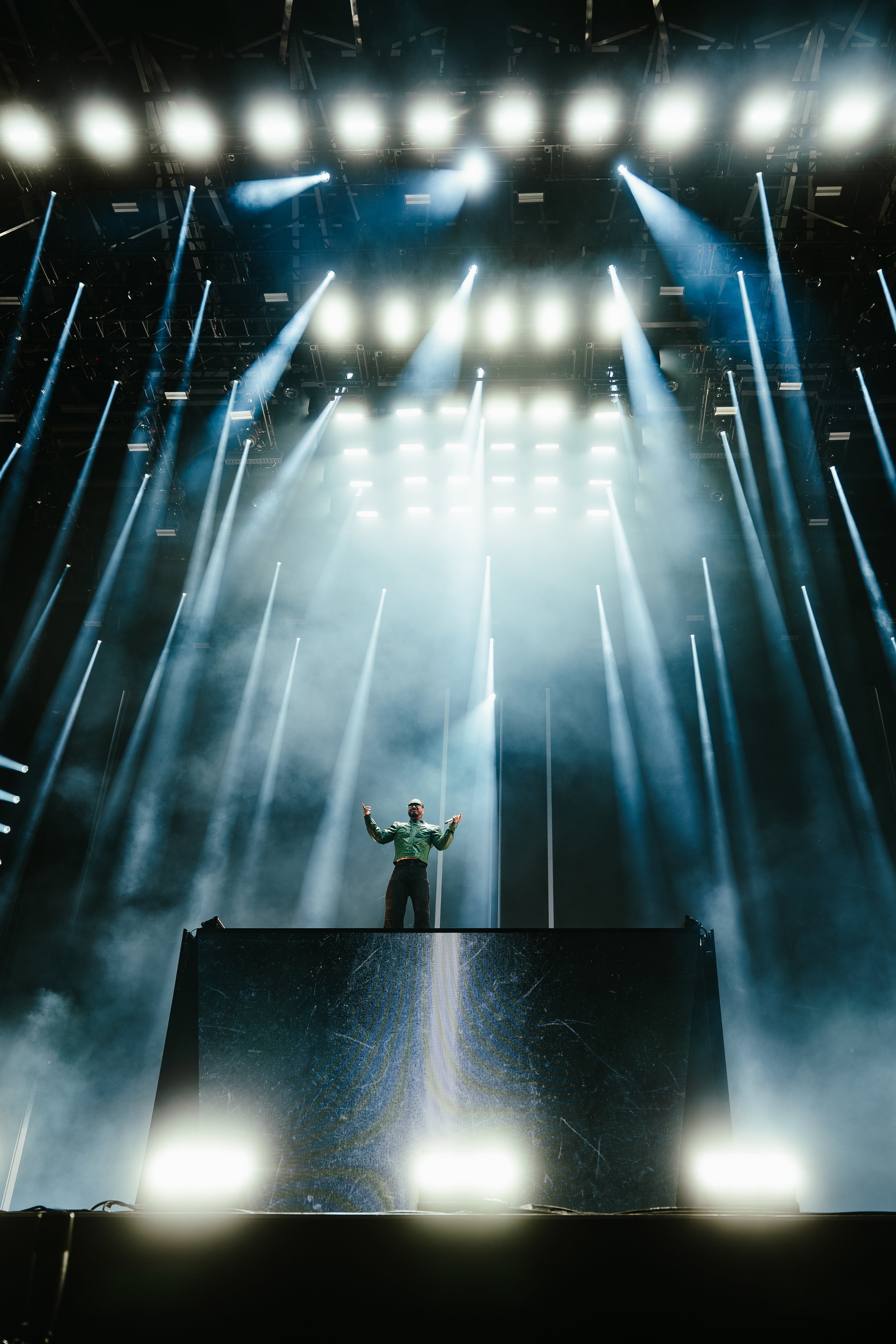 A performer on a stage with bright lights and fog, standing behind a DJ booth or platform. JBalvin FEQ