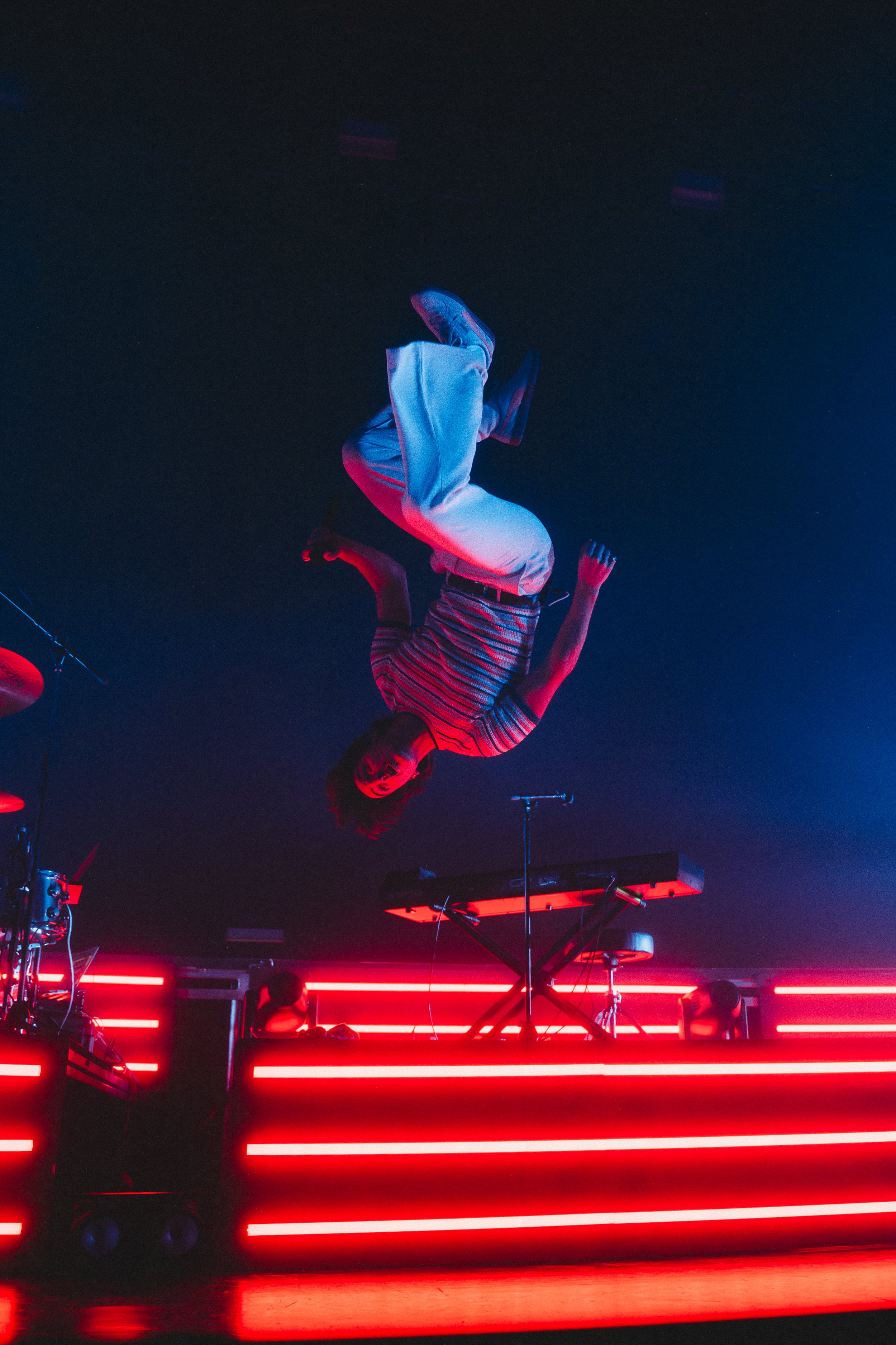 A man performs a dance move with his body upside down over a stage lit with red neon lights, with musical equipment including a keyboard and drums.