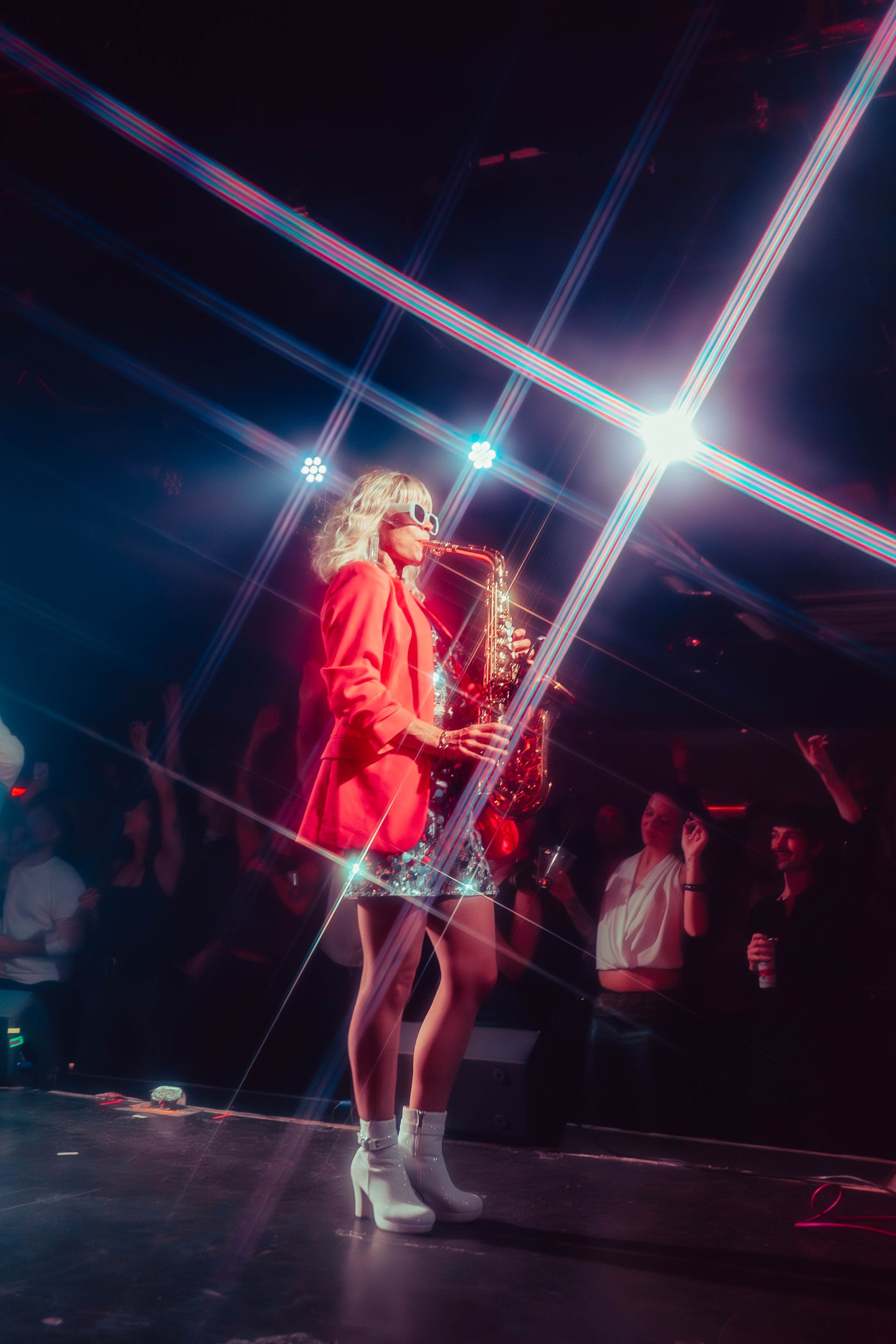 A woman with blonde hair, wearing white sunglasses, a red blazer, and white high-heeled boots, playing a saxophone on stage under bright, colorful lights with an audience in the background.