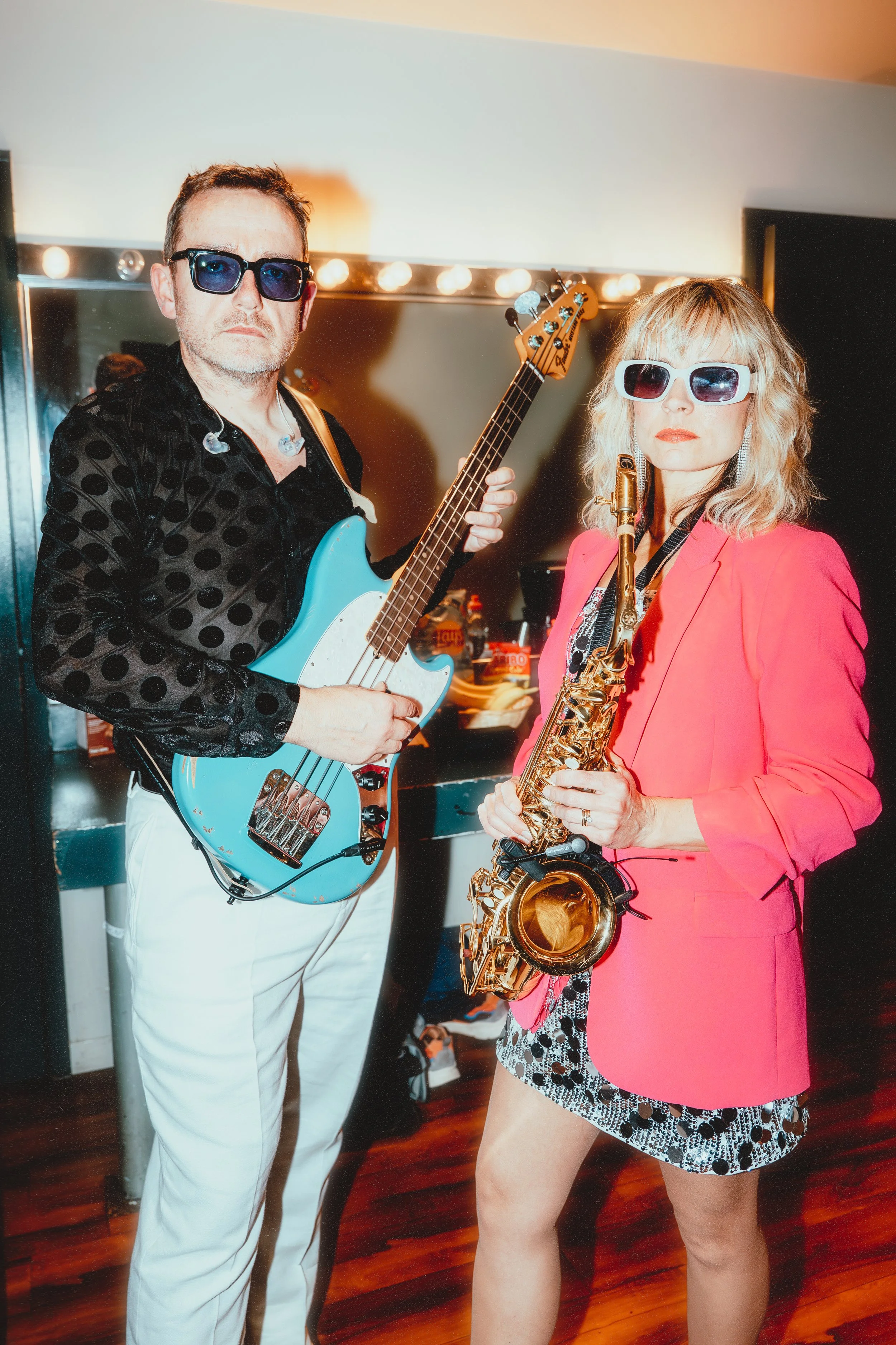 Man and woman with musical instruments, the man with a blue electric bass guitar and the woman with a gold saxophone, both wearing sunglasses and vibrant clothing, standing in a music studio.