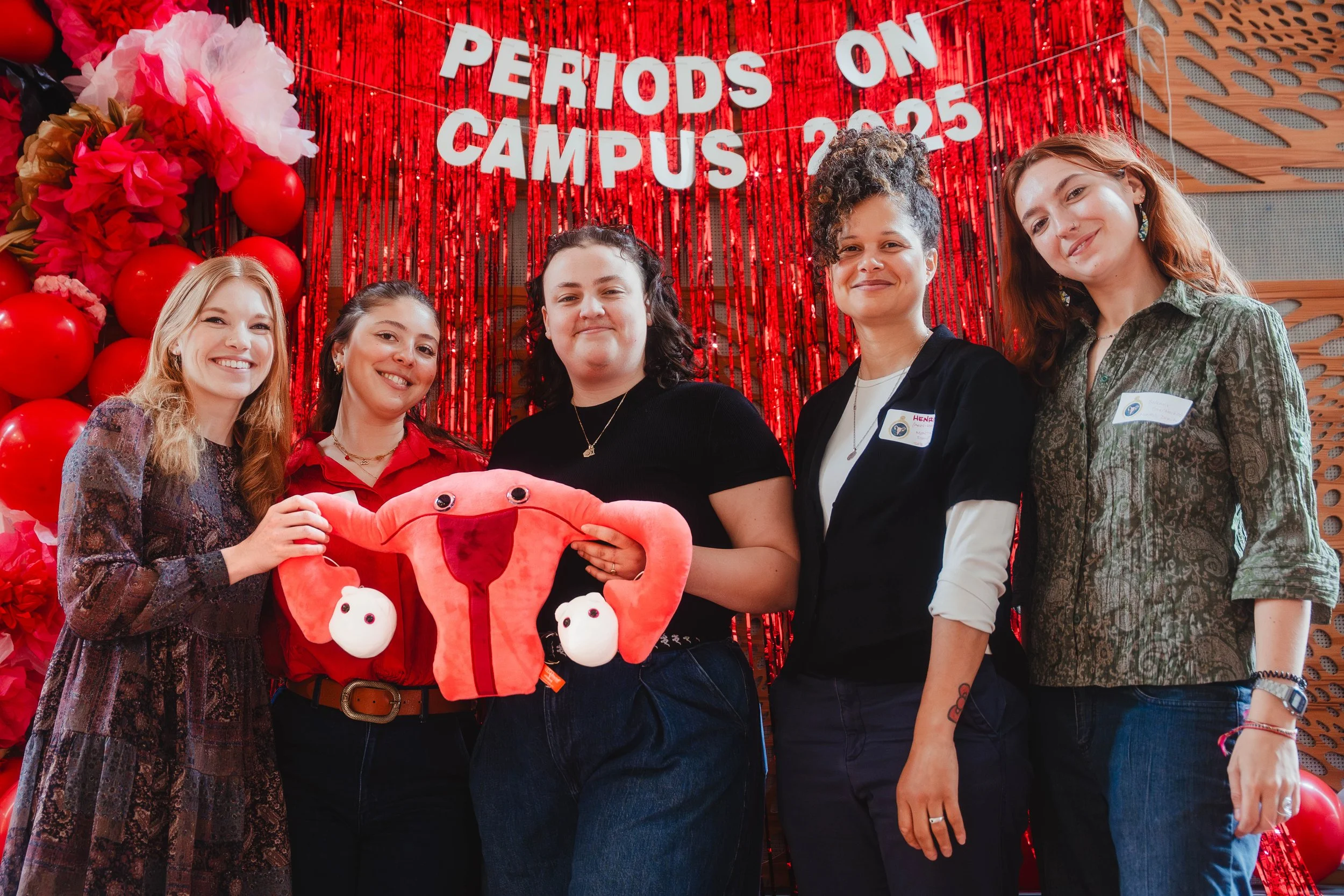 Group of five young women standing together at an event, holding a plush uterus with ovaries, in front of a red backdrop with balloons and a sign that reads 'Periods on Campus 2023'.