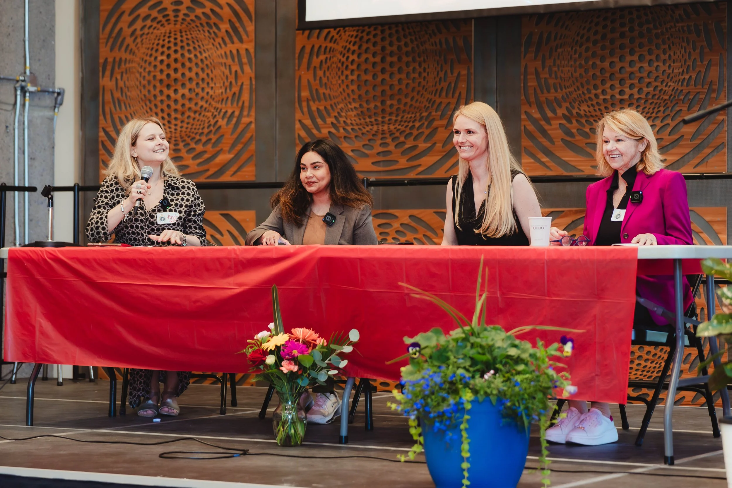 Four women sitting at a panel table with a red tablecloth, on stage, with one woman speaking into a microphone. There are flowers in vases on the floor in front of the table, and a decorative wooden wall behind them.