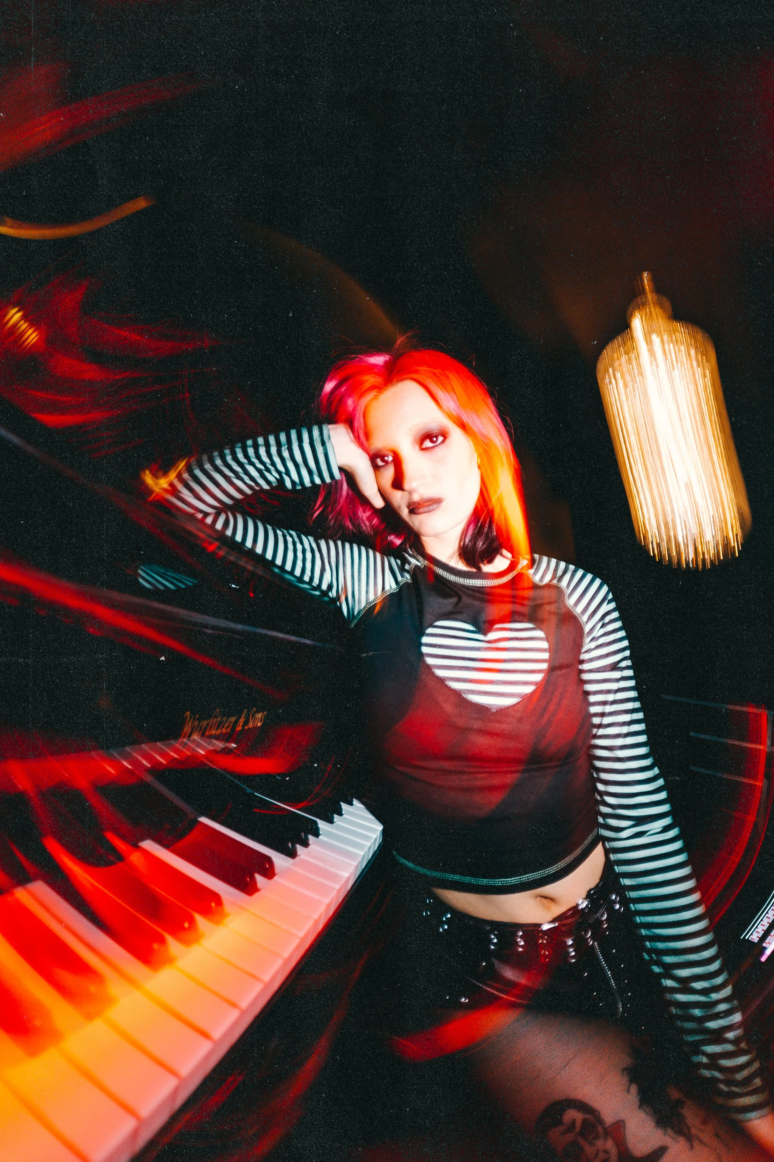 A woman with red and black hair, wearing a black top with striped sleeves and a heart design, poses next to a piano in a dimly lit room with a chandelier. Grae