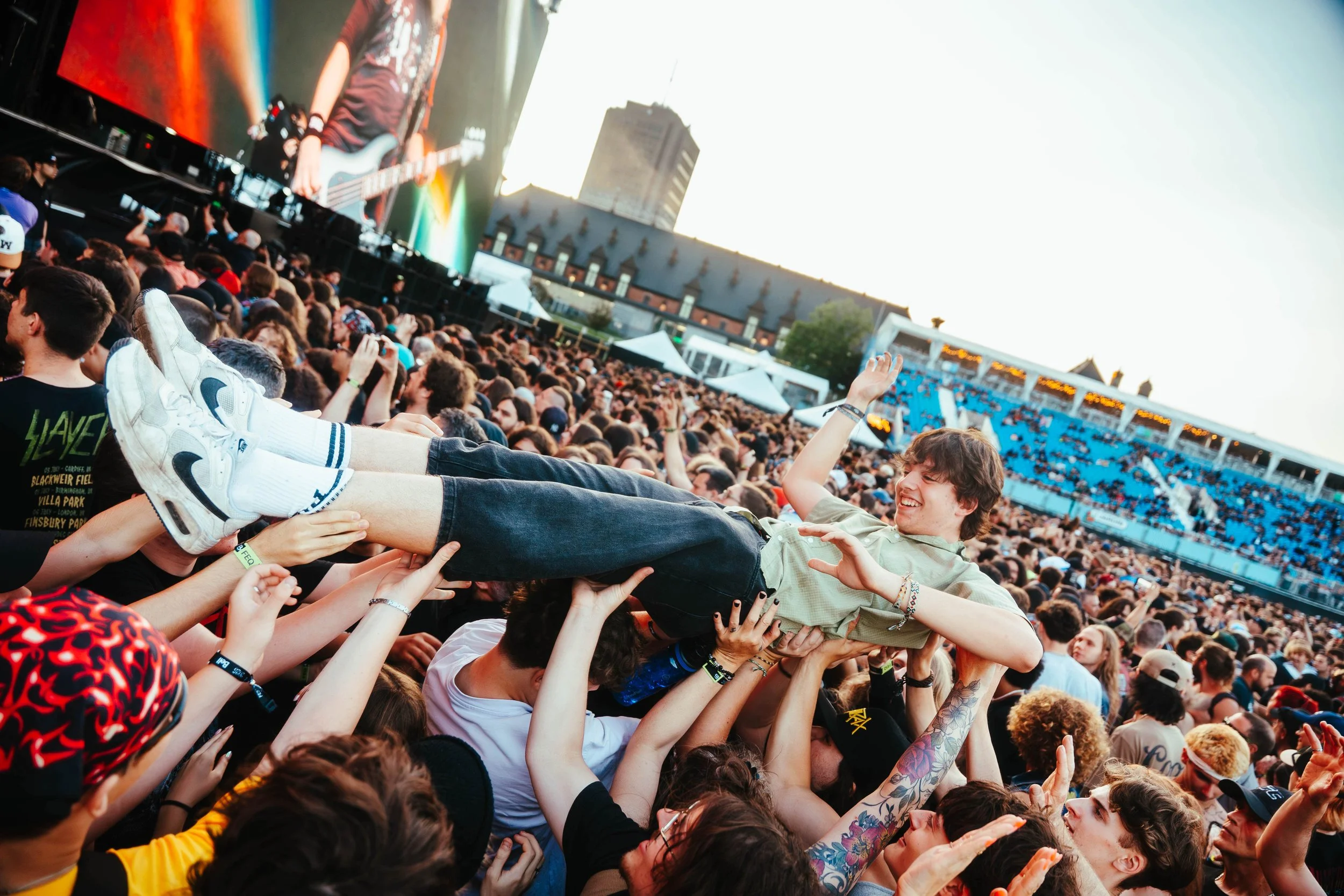 A crowd at a concert or music festival holding up a young man in the air, who is smiling and waving. The large stage with a guitarist and an outdoor venue with seats and tents are visible in the background. FEQ Slayer