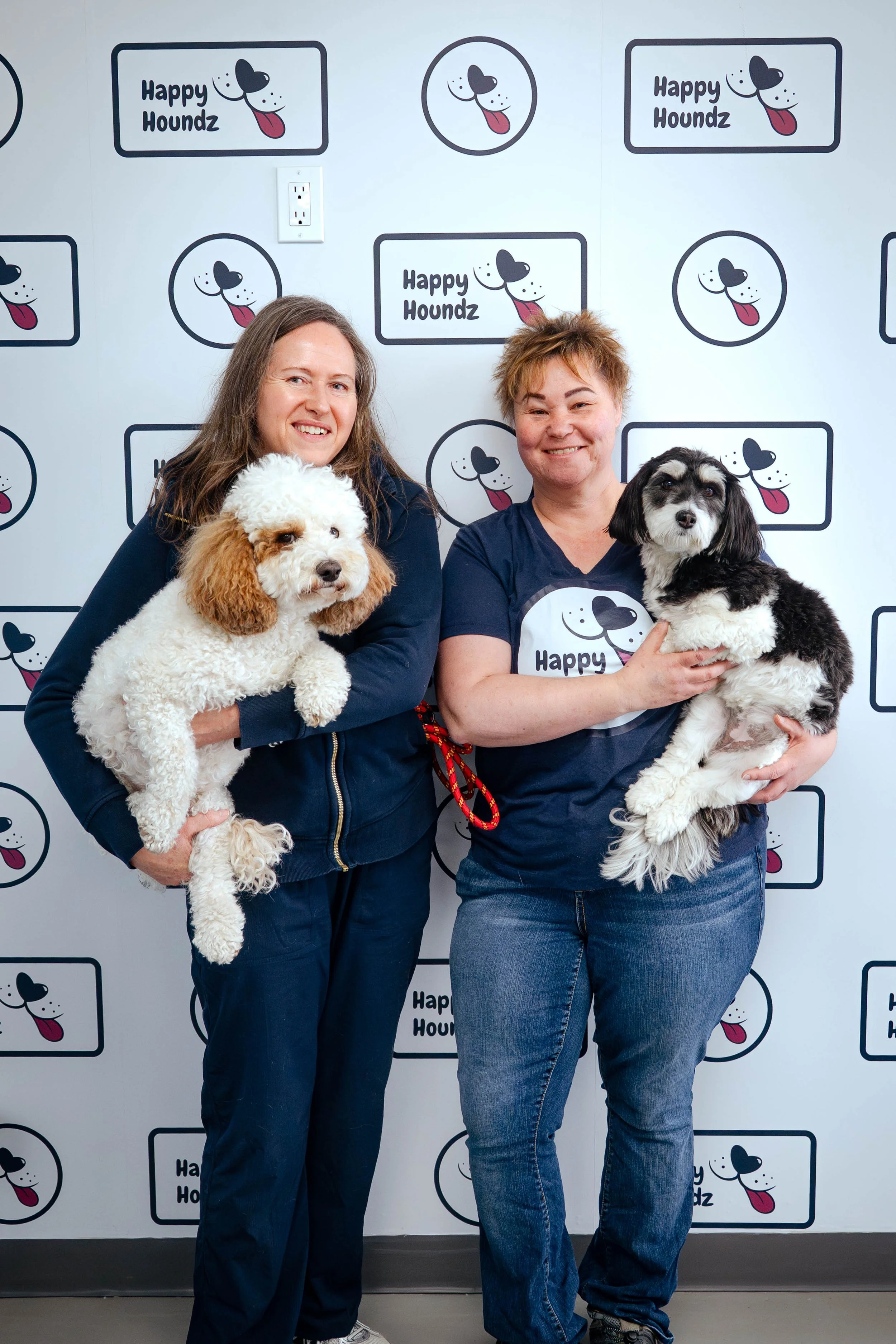 Two women holding dogs and smiling in front of a backdrop with Happy Houndz logos featuring cartoon dog faces with tongues out.