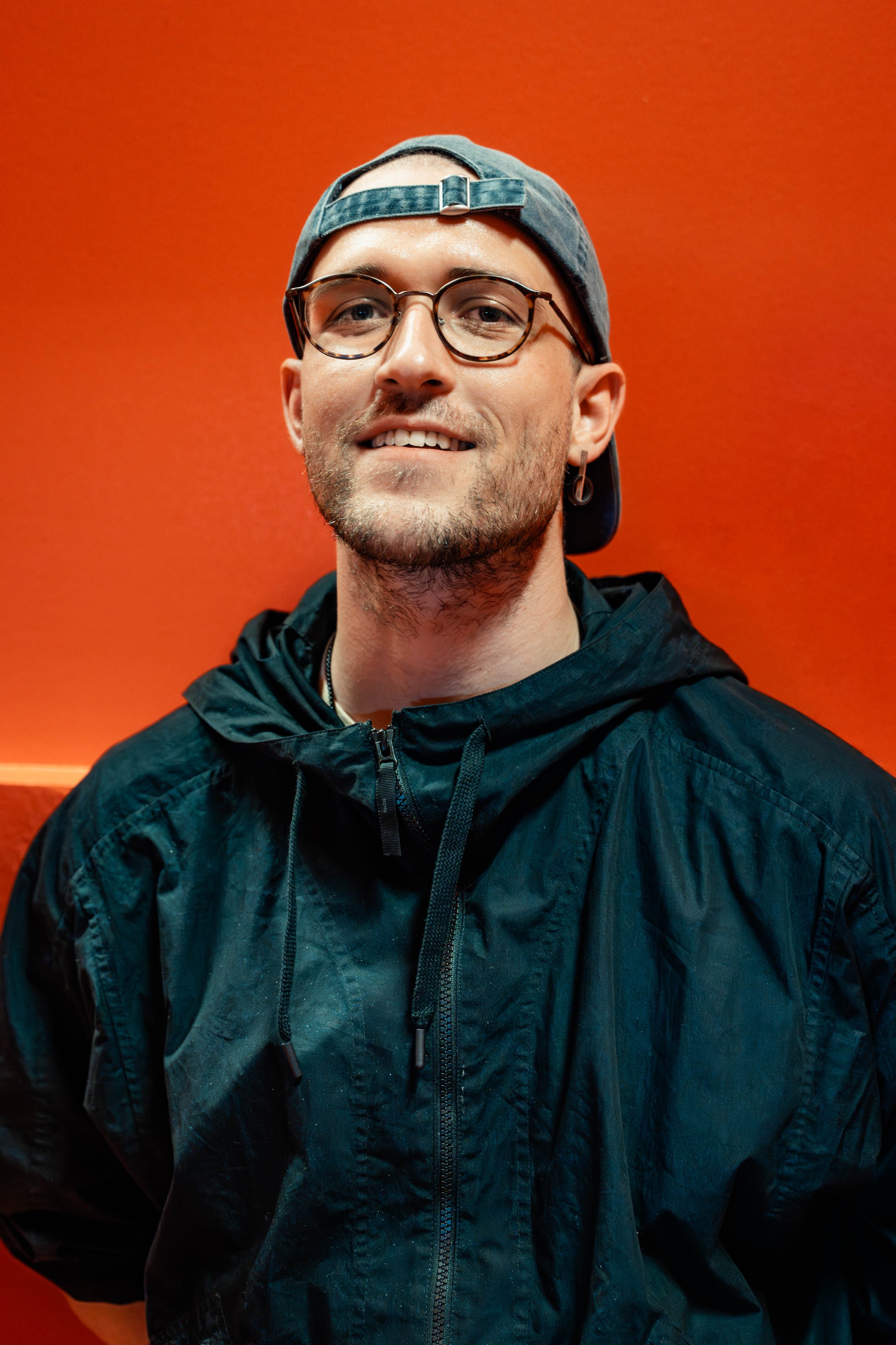 A young man wearing glasses, a grey cap backward, and a black jacket, smiling in front of an orange wall.