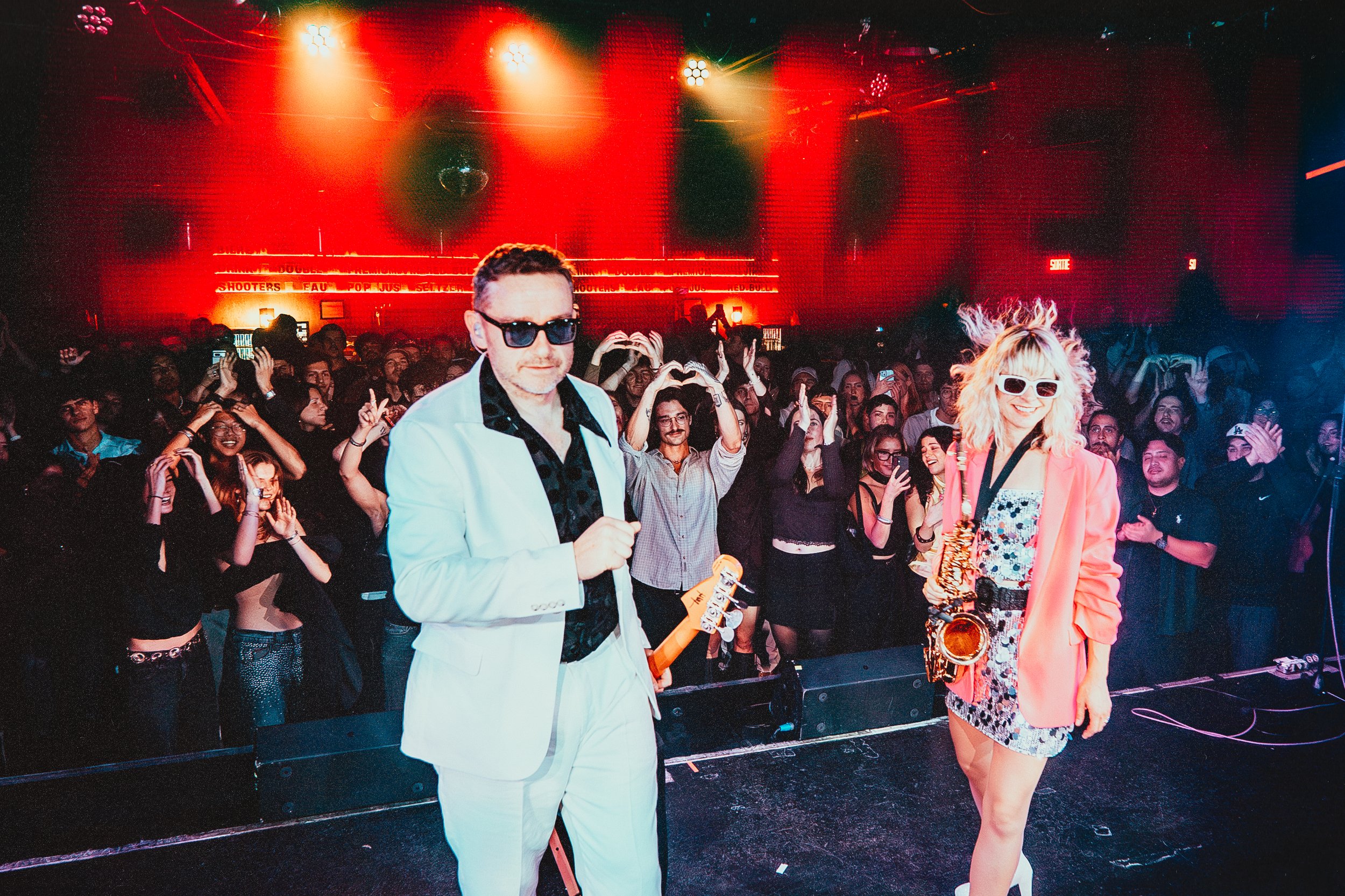 Two performers on stage with a crowd of people in a concert or music festival setting, colorful lighting, one male and one female, both wearing sunglasses, the male in a white suit and the female in a bright pink blazer and floral dress, with people 