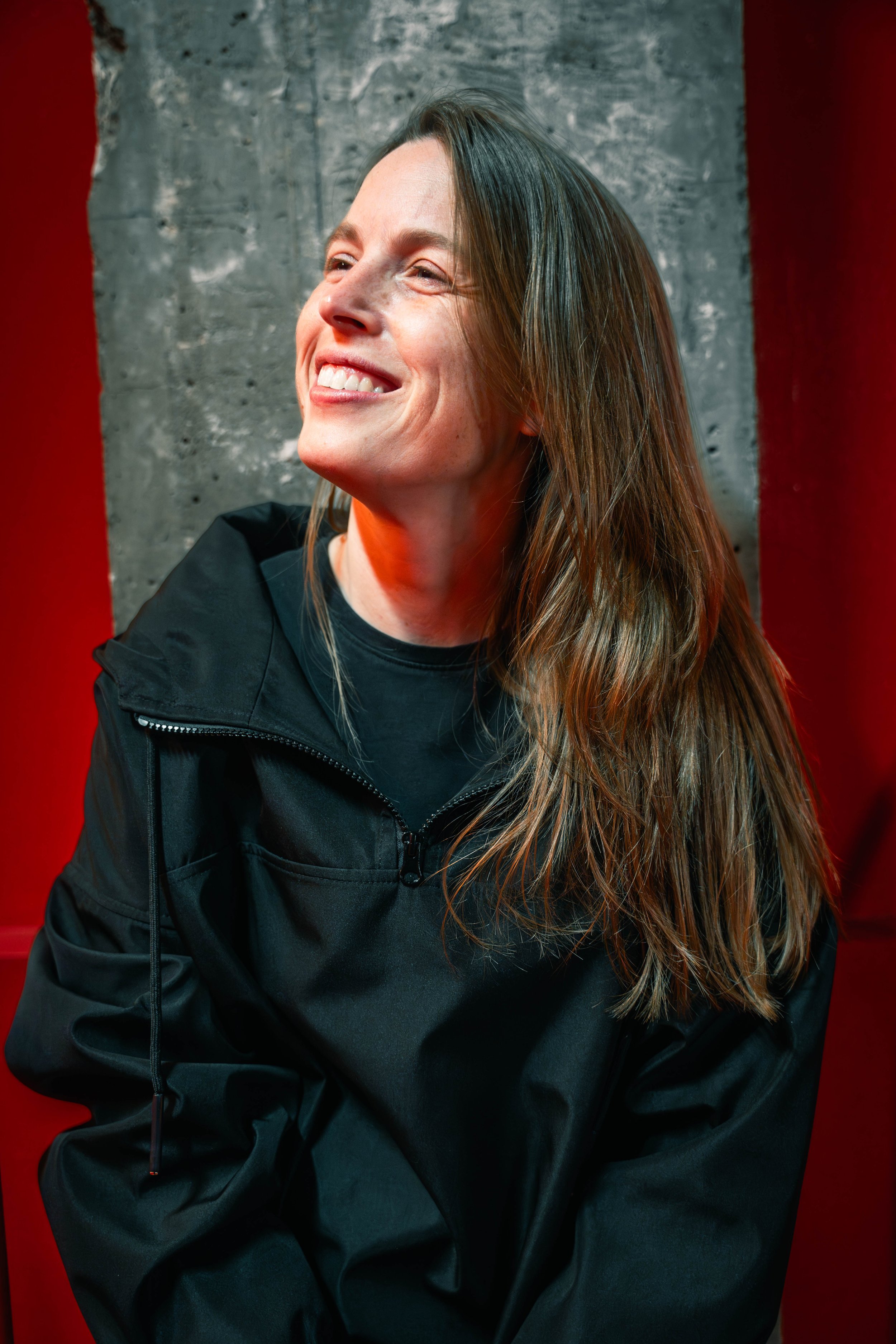 A woman with long brown hair smiling, wearing a black jacket, in front of a gray concrete wall and red background.