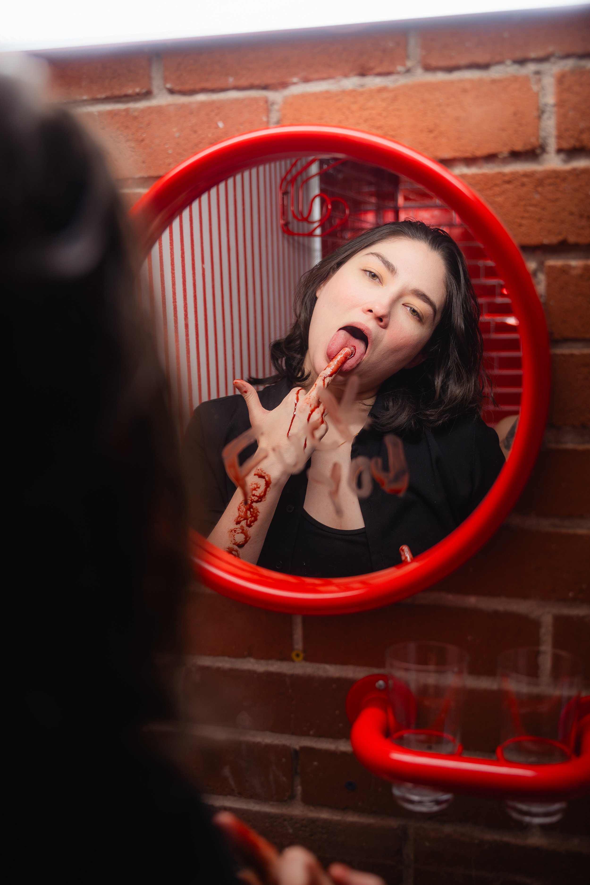A woman with dark hair looking into a mirror, licking her finger which has red paint or substance on it, with some of it on her hand. She is in front of a red frame mirror mounted on a brick wall.