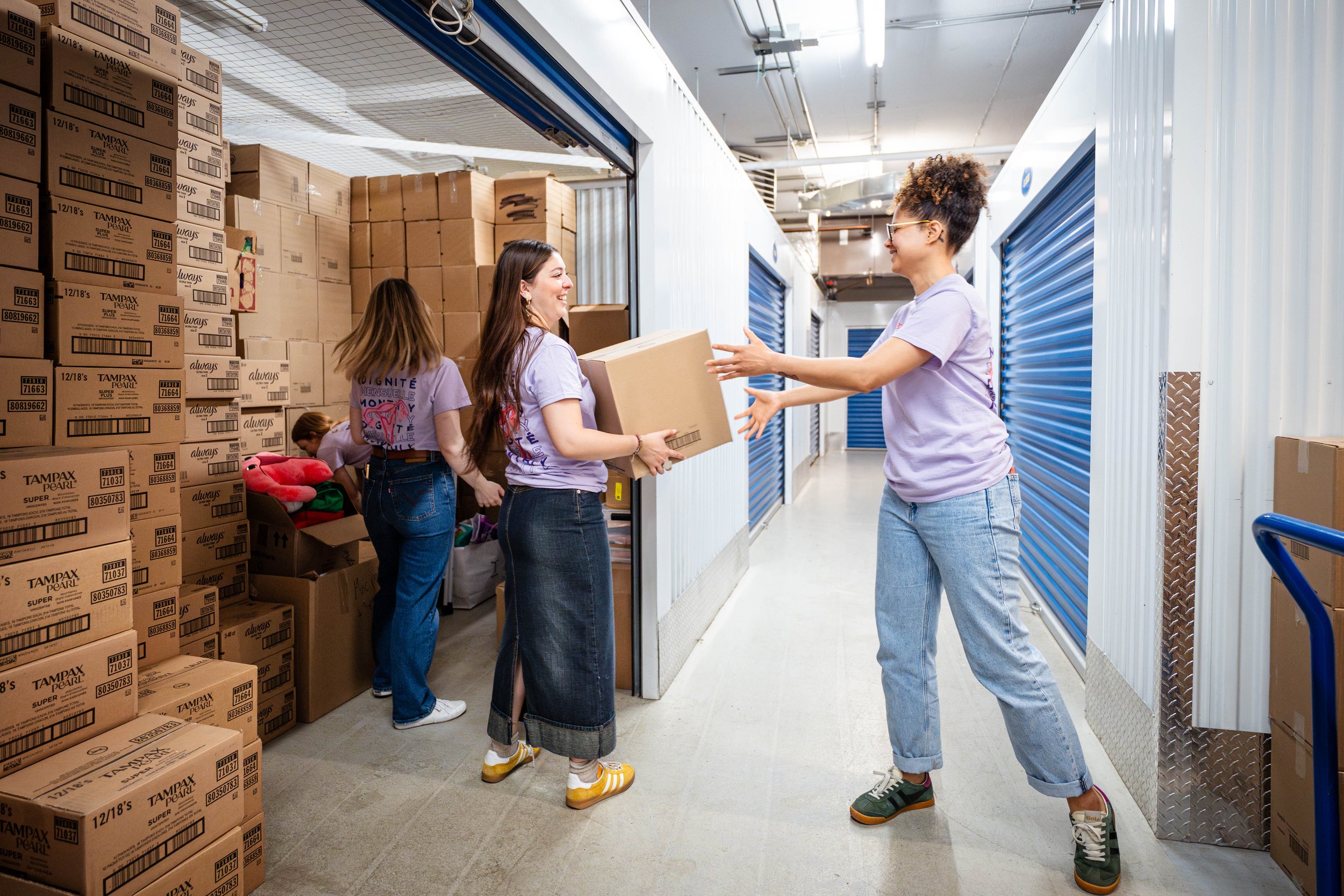 People working in a storage unit, with one person handing a box to another, smiling, surrounded by packed boxes and storage units.