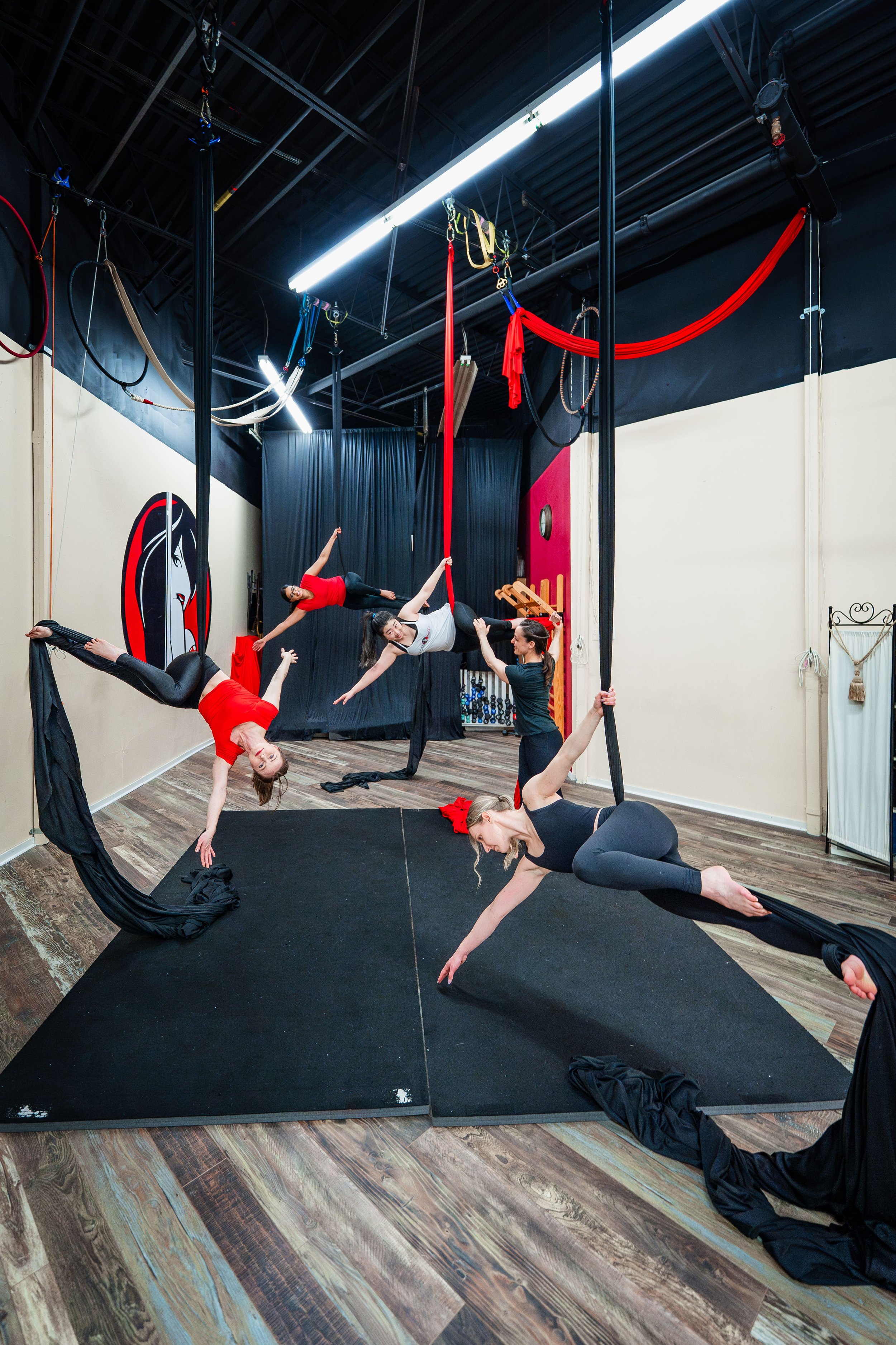 Four women practicing aerial acrobatics with silks and aerial hoop in a circus studio, with black curtains, wood flooring, and circus equipment hanging from the ceiling.